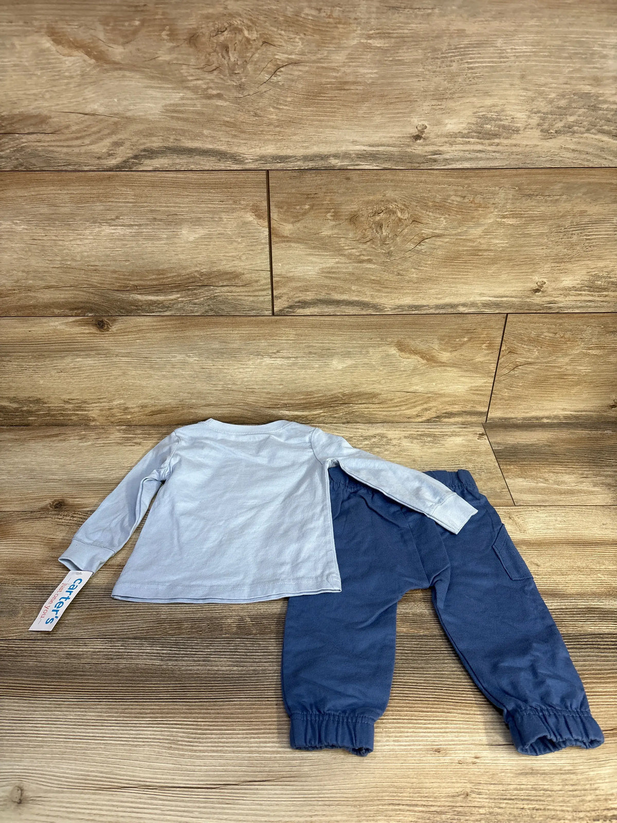 Top-down view of Just One You 2-piece blue shirt and pants set in size 9 month, showing long sleeves and elastic cuffs on wood flooring
