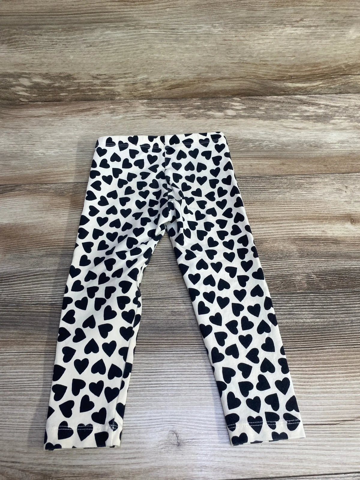 Flat lay of Tea Collection Heart Print Leggings in ivory showcasing black heart print all over on wood plank floor