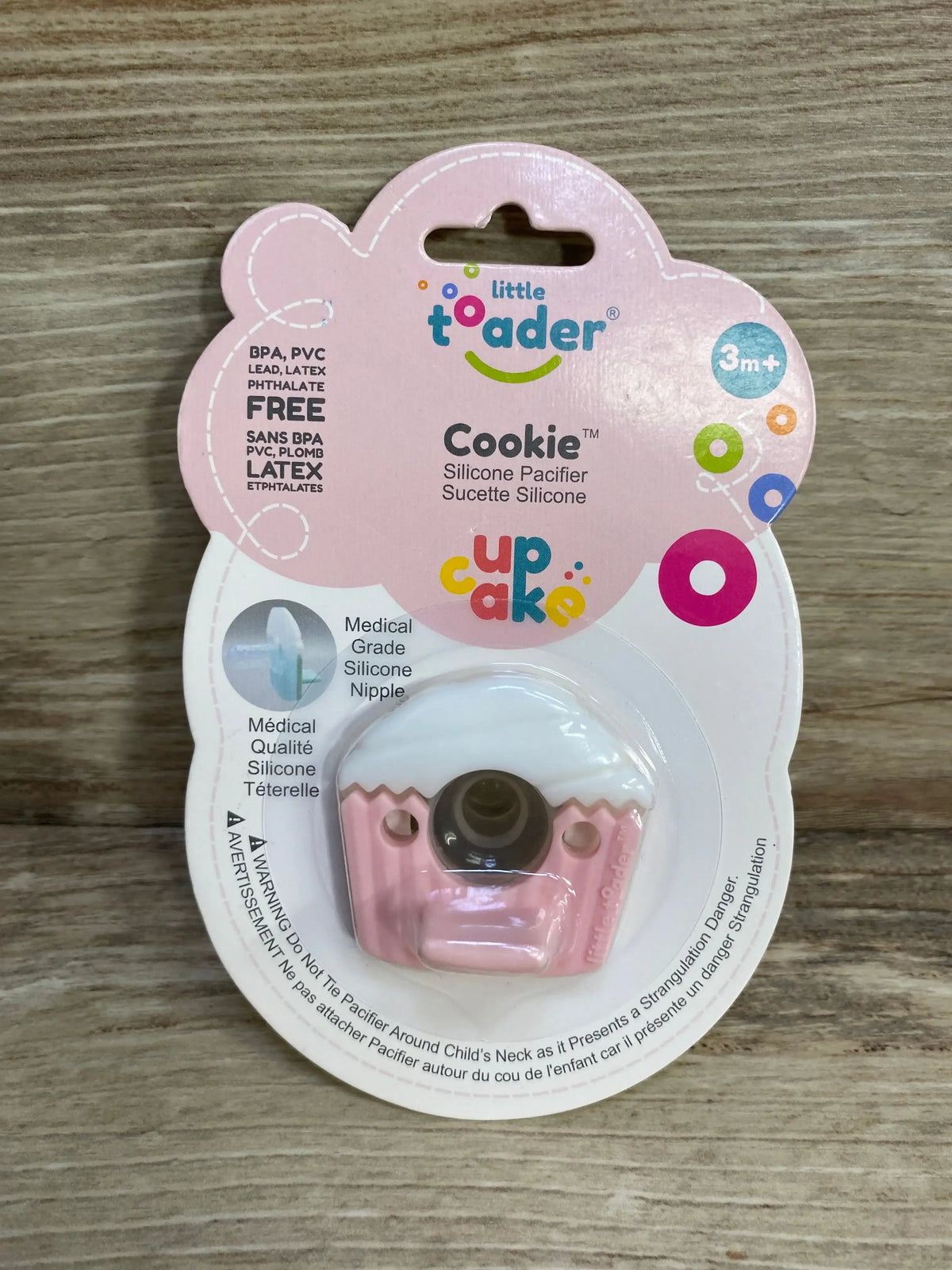 Front view of Little Toader Cookie cupcake silicone pacifier in pink in original blister packaging showing 3m+ age label on wood surface