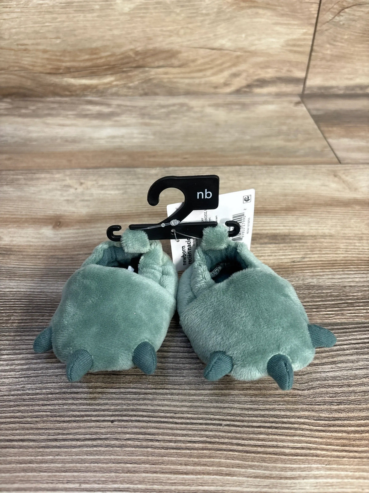 Child Of Mine plush green dinosaur claw slippers for infants on a wooden surface.
