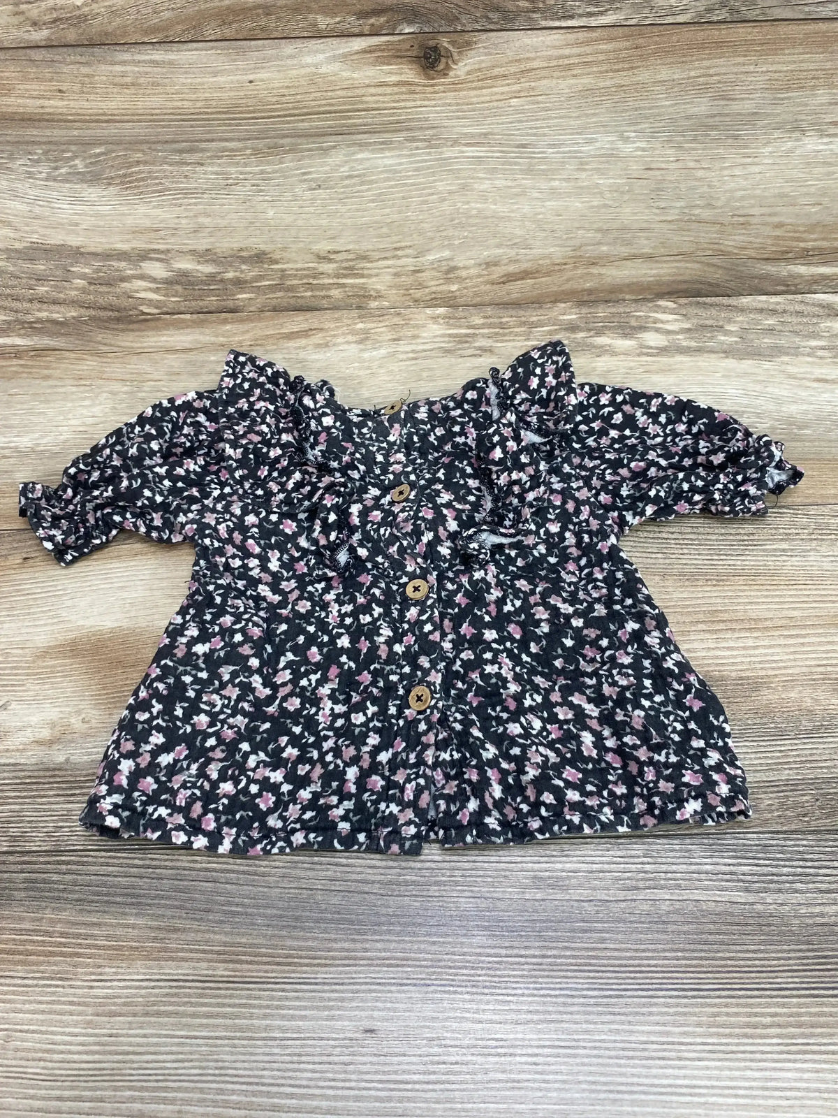 Front view of Jessica Simpson black floral ruffle top, size 12m, showcasing ruffled sleeves and button front on wood grain