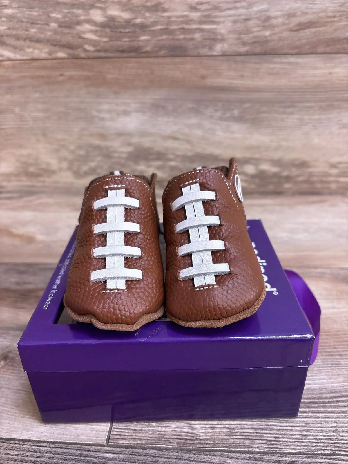 Brown baby shoes designed like footballs with white laces, placed on a purple box on wooden surface