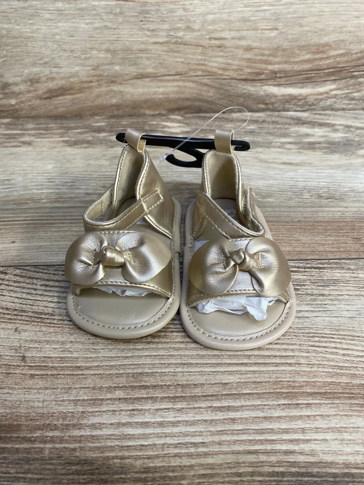 Front view of baby gold Child of Mine sandals showcasing knotted bow straps and Velcro ankle closure on wood-look background