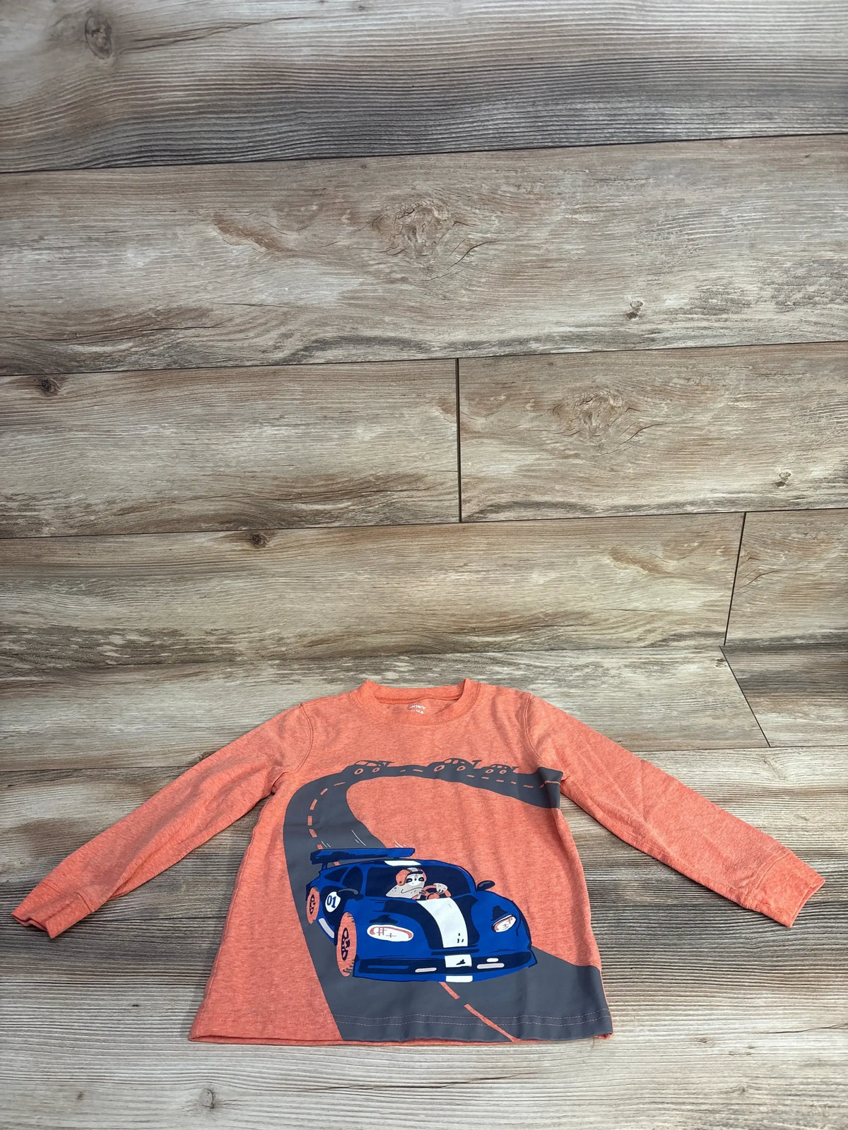 Orange long-sleeve kids shirt with blue race car and dog driver graphic on wood floor