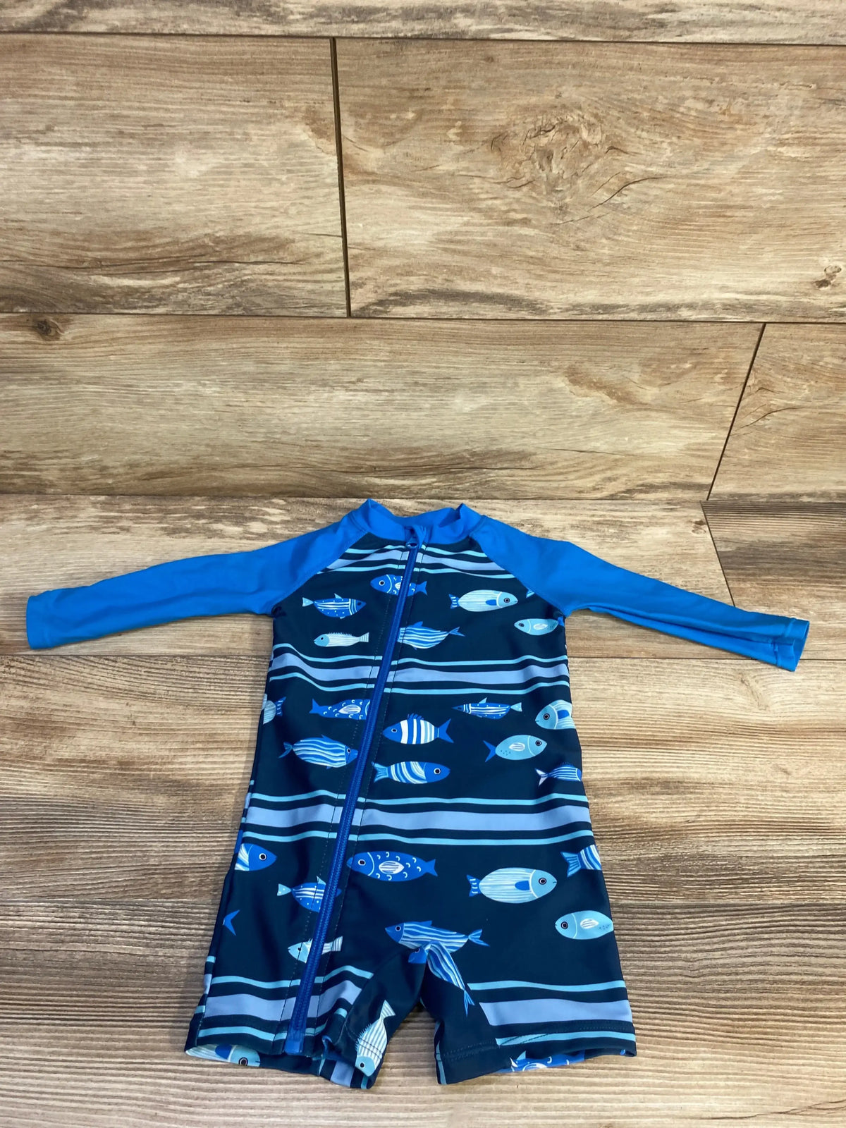 Flat lay of baby long-sleeve 1-piece rashguard swimsuit size 3-6M showing front zipper and blue fish print on wood background
