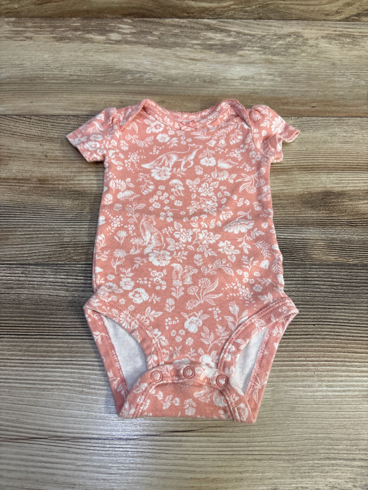 Top-down view of Carter’s pink floral short-sleeve baby bodysuit showcasing white floral print and three snap closures on wood background