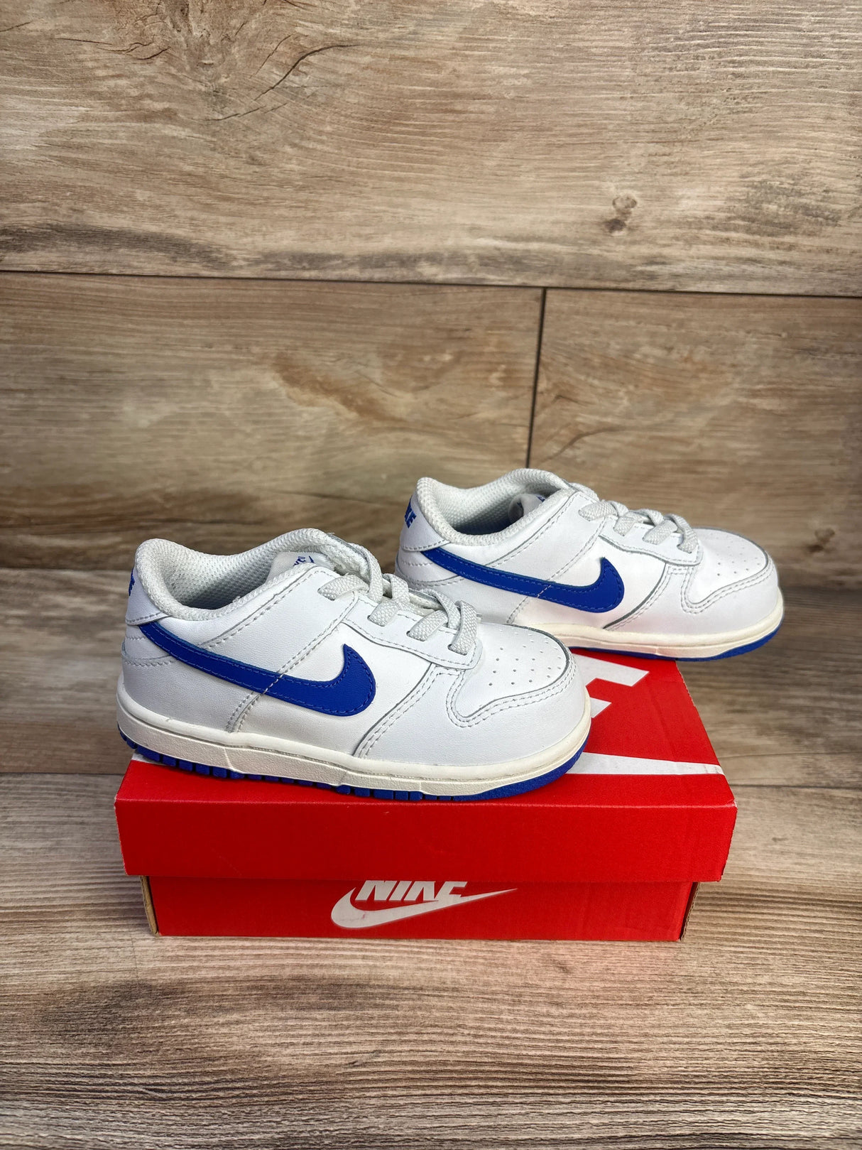 Side view of Nike Dunk Low toddler sneakers, white leather with blue Swoosh and blue sole, on a red Nike box on wood background