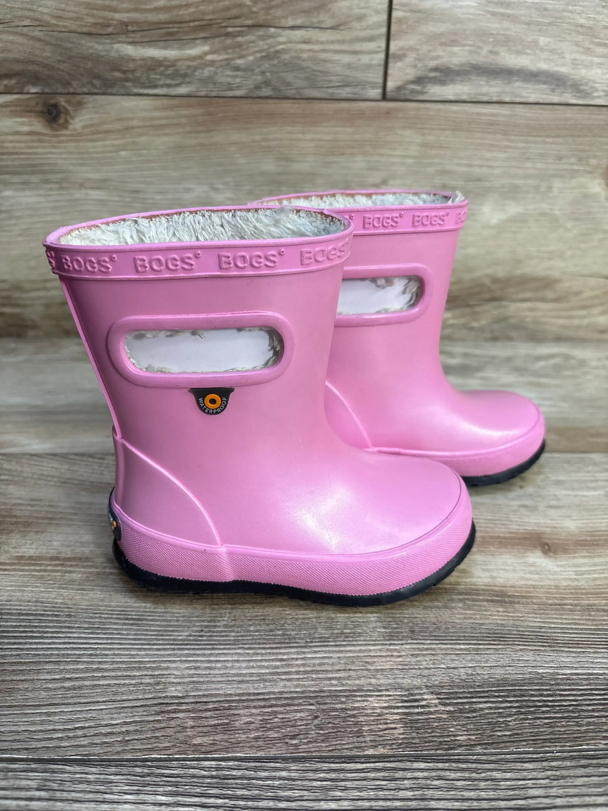 Side view of Bogs Skipper pink toddler rain boots showing carry handles and fleece lining on wood floor