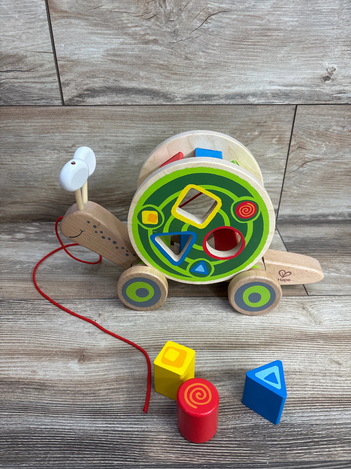 Wooden snail pull toy with green patterned shell and colorful shape sorter blocks on wooden floor