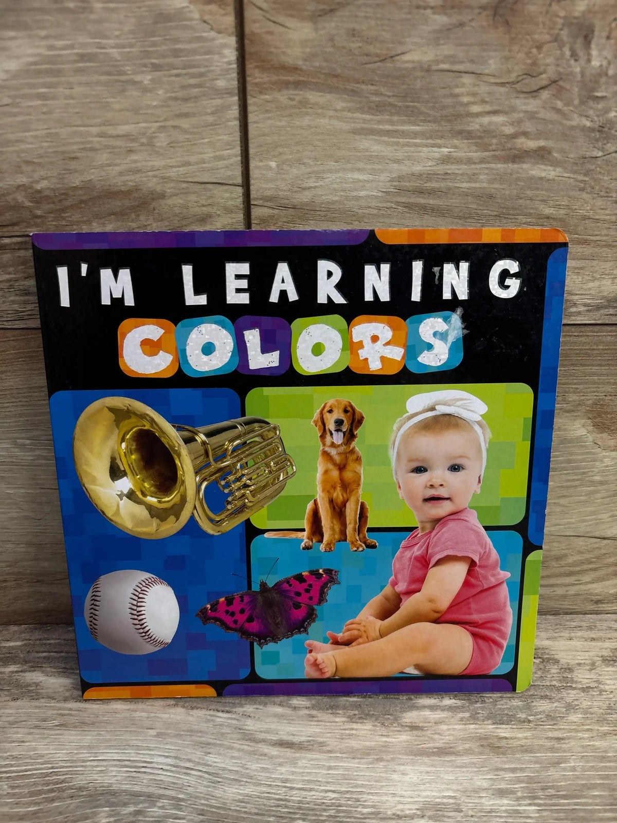 Front of I'm Learning Colors board book showing a colorful cover with baby in pink onesie, dog, tuba, and butterfly on wood