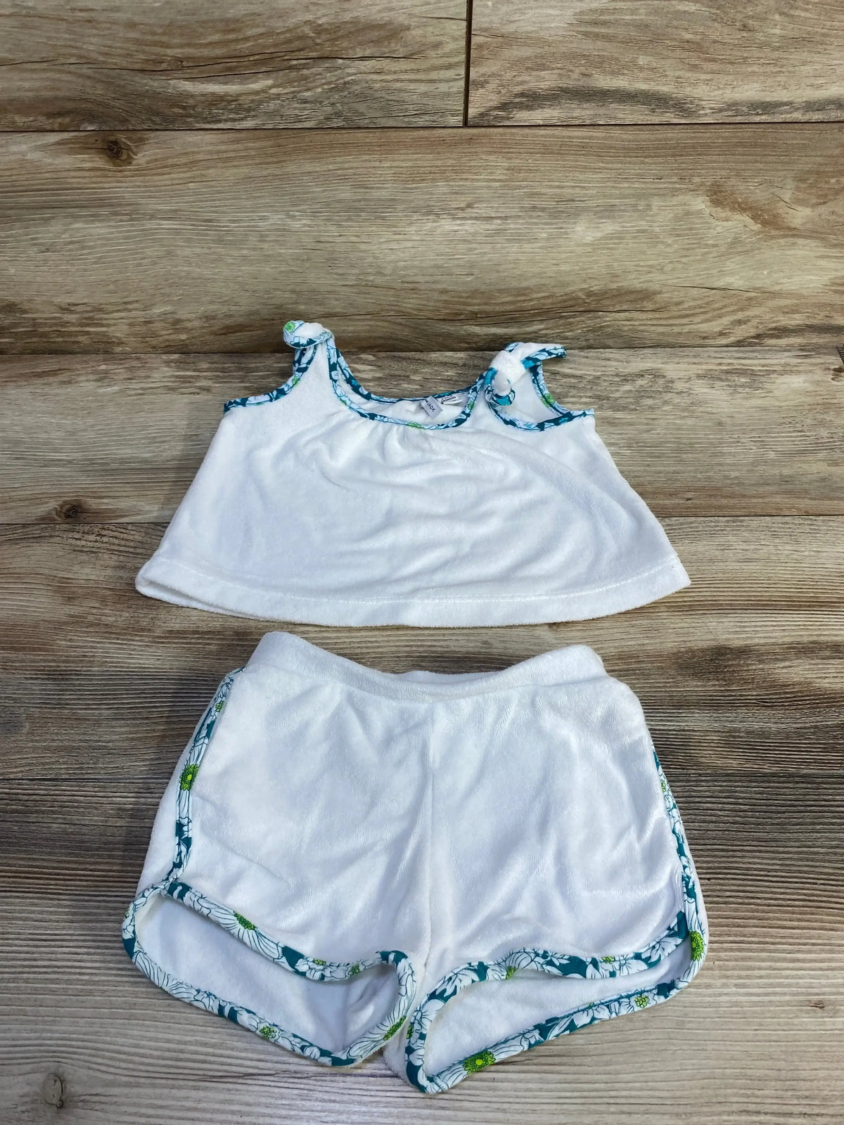 Flat lay of white terry tank top and shorts showing teal floral trim and knotted shoulder straps on light wood background