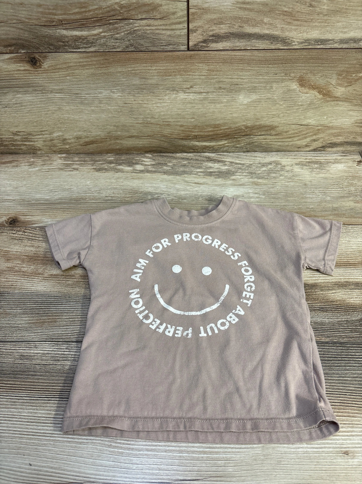 Flat lay of Grayson mini shirt in brown with white smiley and circular text ‘Aim for progress forget perfection’ on wood