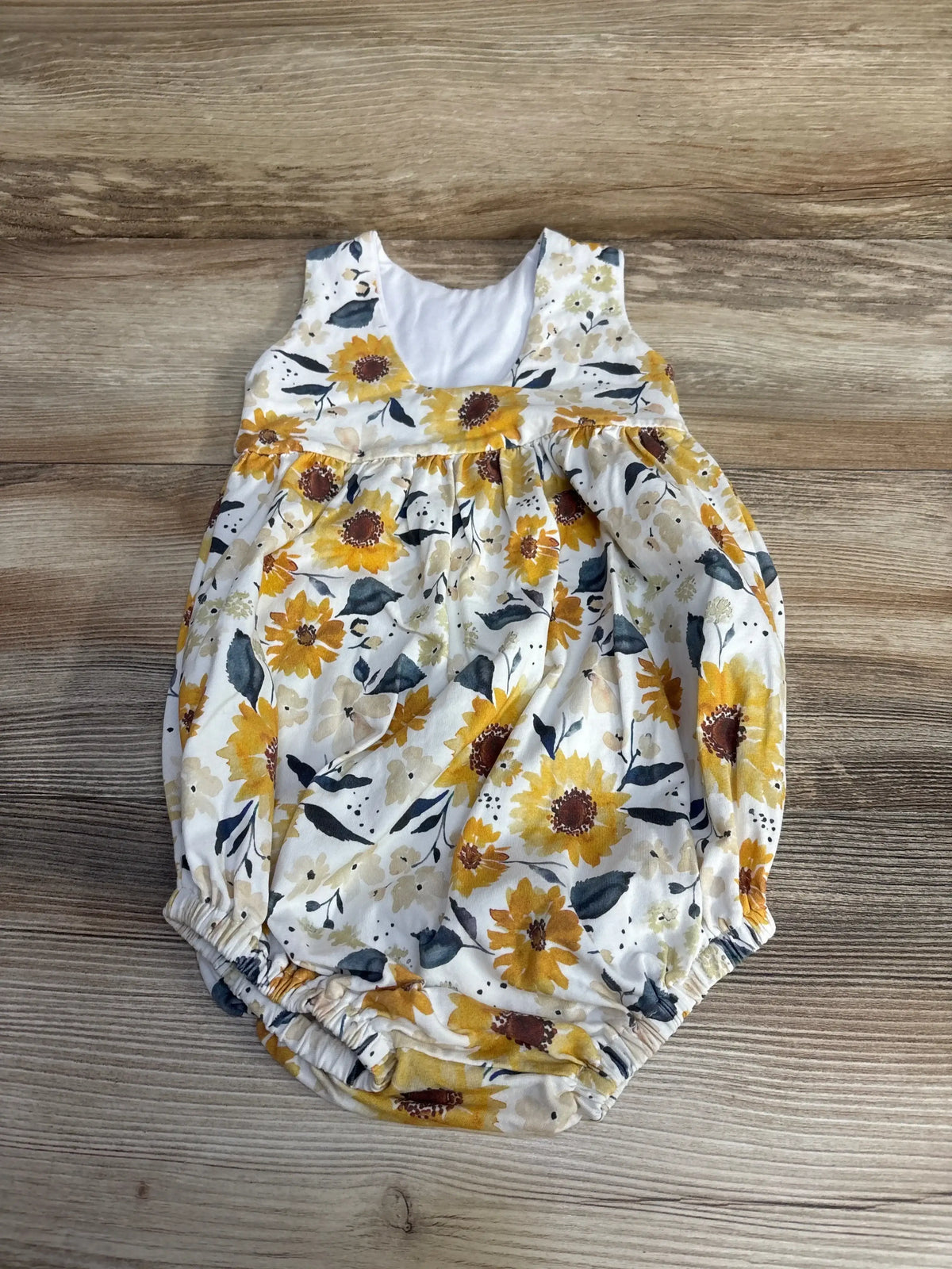Top-down view of white handmade floral romper (4T) showcasing yellow sunflower print and elastic leg openings on wood background