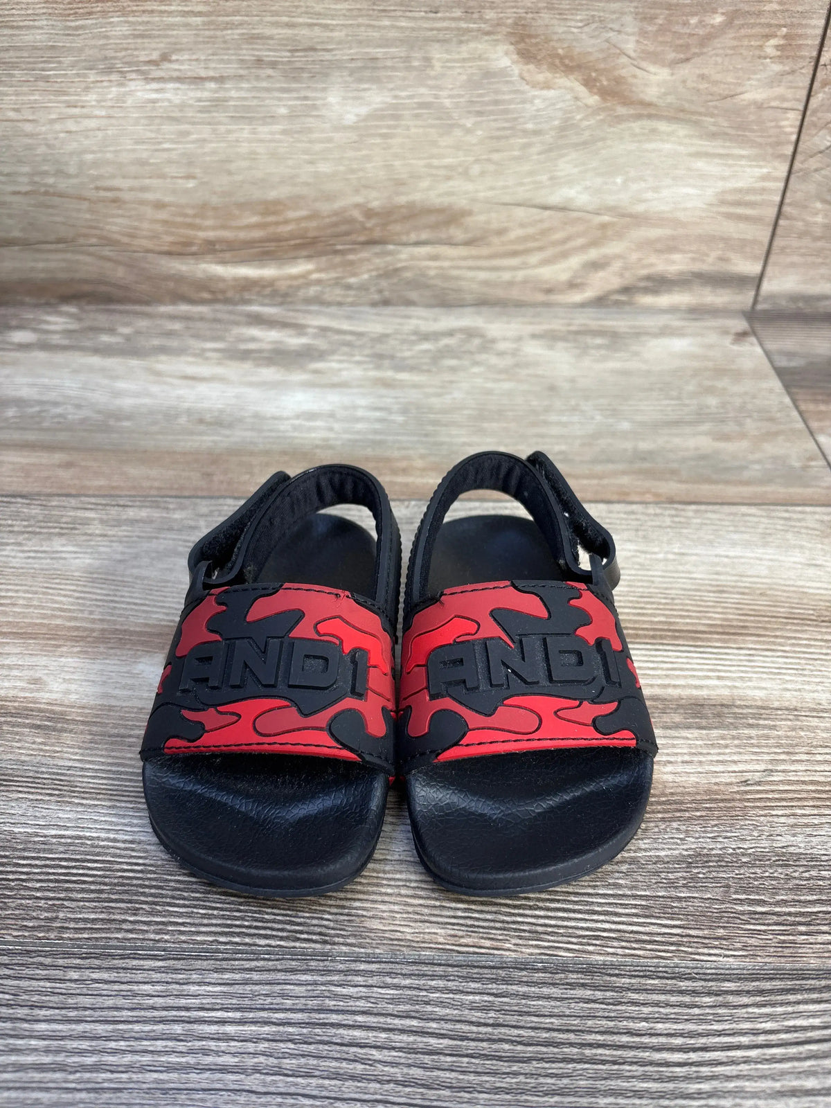 Front view of And1 camo slide sandals showcasing red and black camouflage straps with black cushioned soles on a wood floor