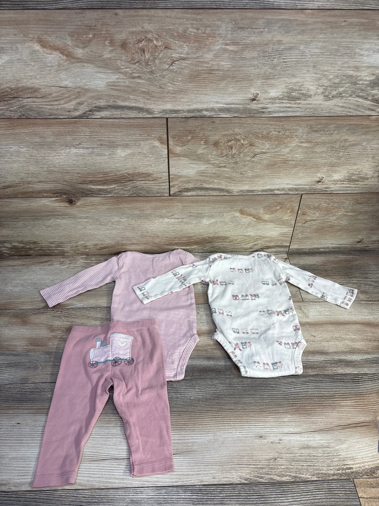 Flat lay of Carter's 3-piece 6M bodysuit set showing a pink striped long-sleeve bodysuit, an ivory long-sleeve bodysuit with small prints, and pink leggings with a train applique on wood backdrop