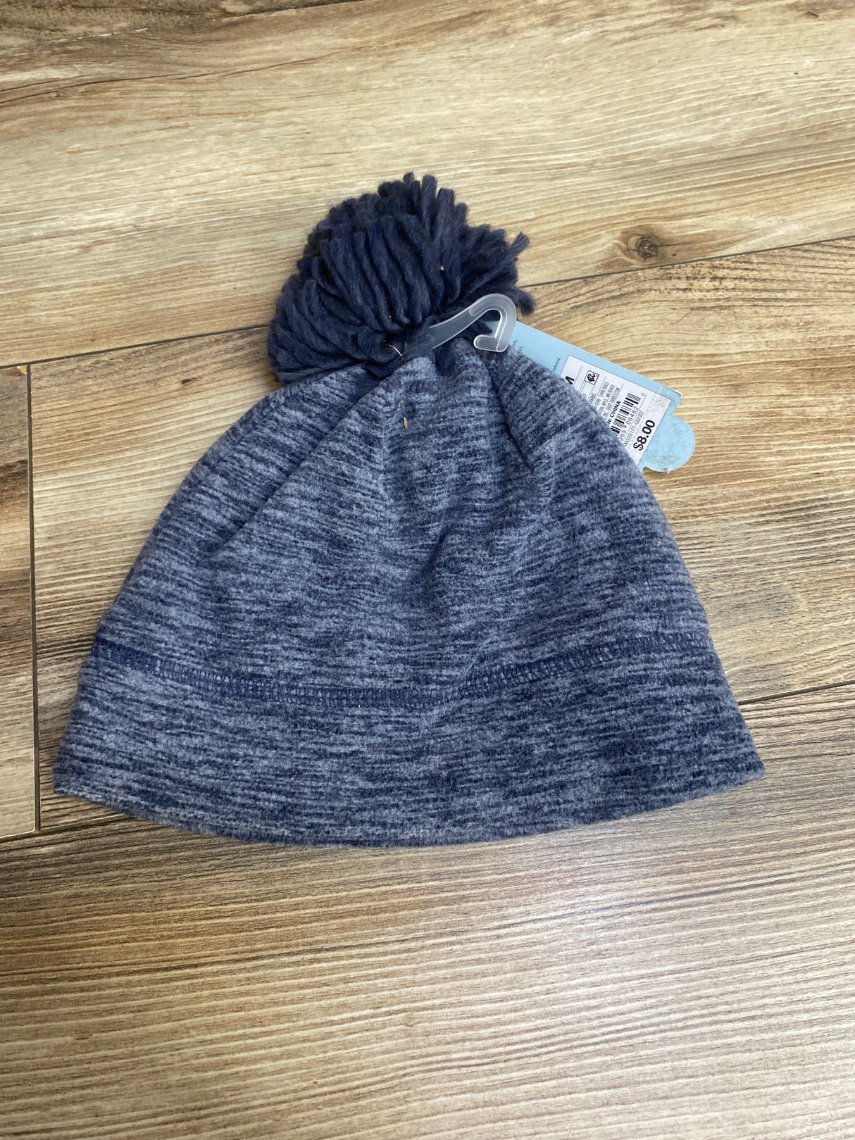 Grey and navy knit winter beanie hat with pom-pom on wooden floor with price tag attached
