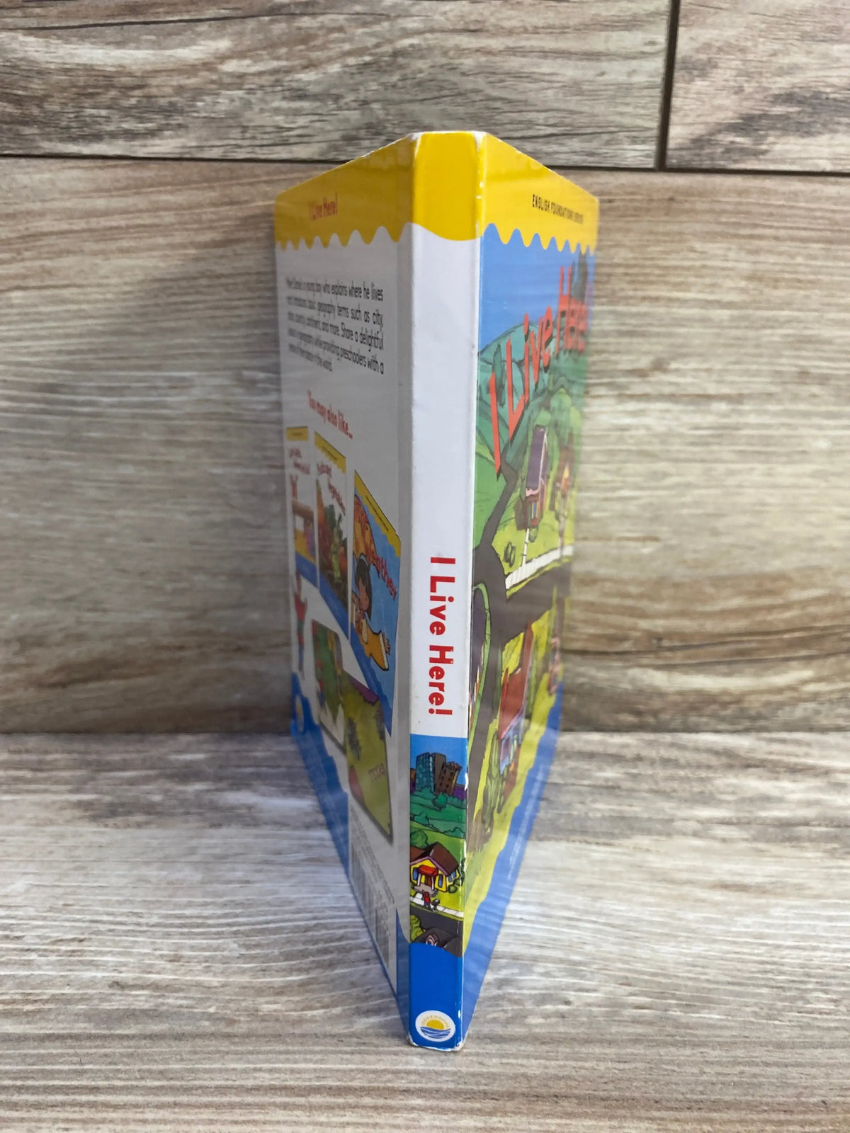 Angled view of I Live Here! board book showing the spine and colorful covers on a wood plank background