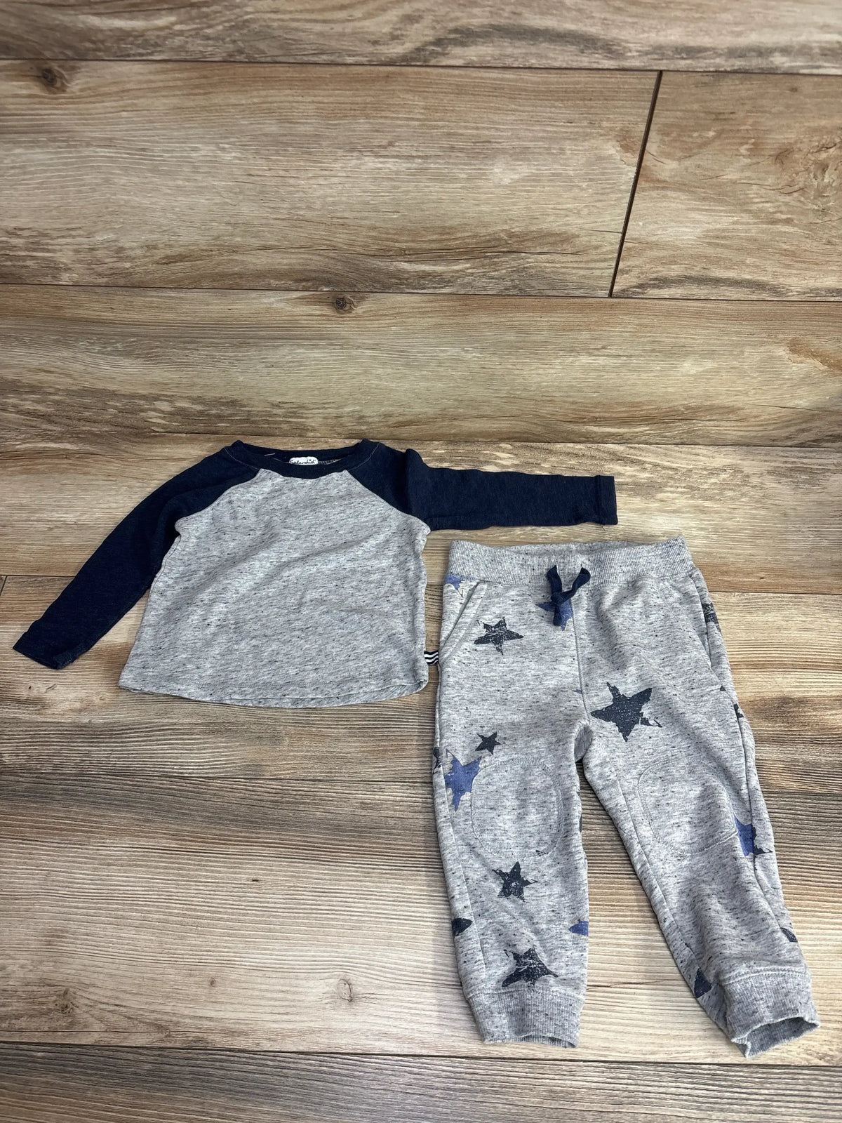 Flat lay of toddler two piece set showcasing grey raglan long sleeve top with navy sleeves and matching heather grey jogger pants with navy star print on wood plank floor