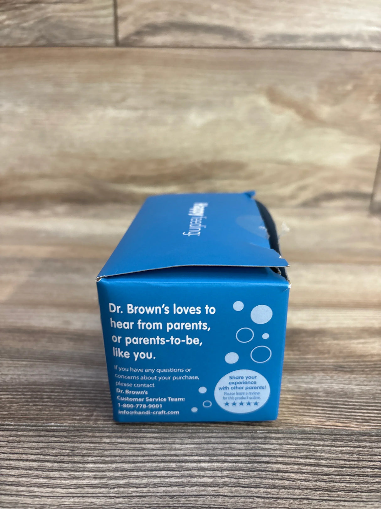 Angled side view of Dr. Brown's Milestones silicone narrow baby bottle handles 3-pack box showing blue packaging with customer message Dr. Brown's loves to hear from parents on wooden tabletop