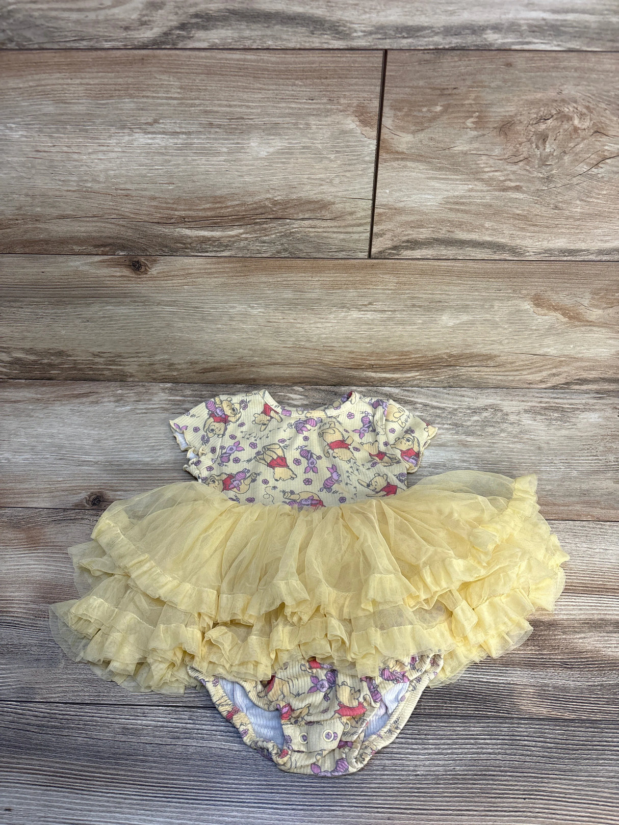 Yellow baby girl romper dress with ruffled tulle skirt and cartoon print, displayed on wood.