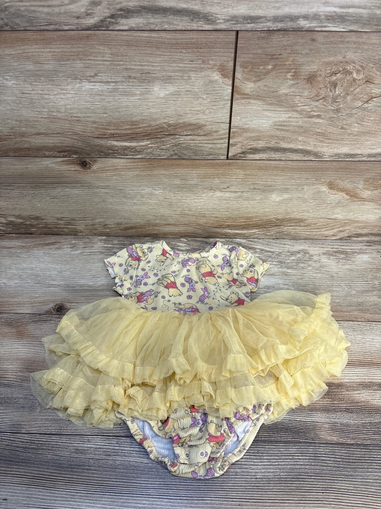 Yellow baby girl romper dress with ruffled tulle skirt and cartoon print, displayed on wood.