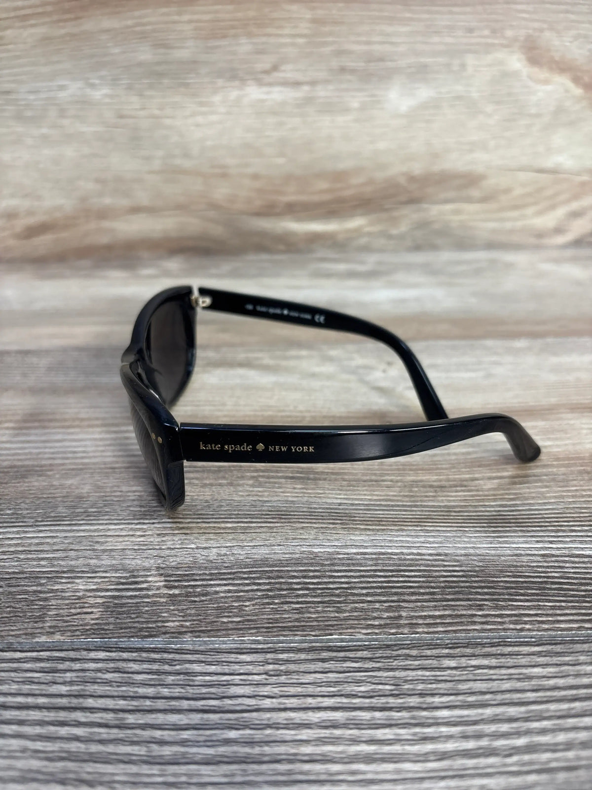 Side view of Kate Spade New York Tamsin/S sunglasses showcasing black rectangular plastic frames with a wood-grain background