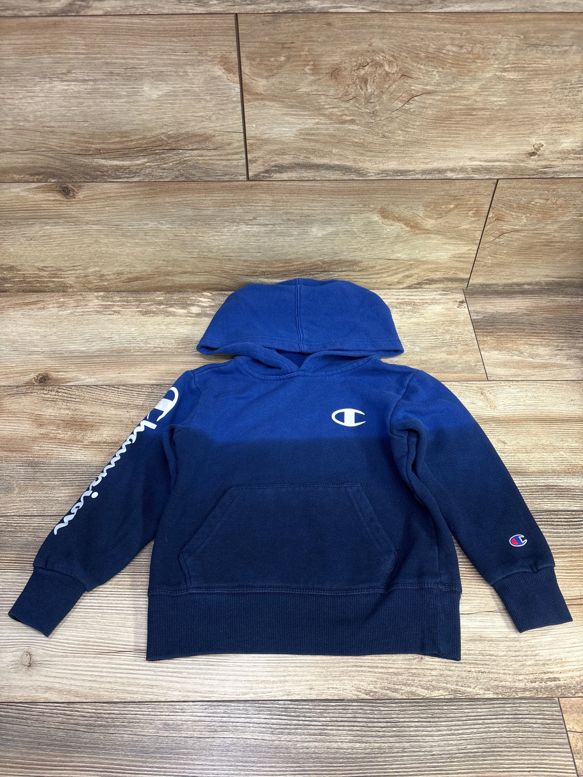 Front view of blue Champion pullover hoodie in size 4T showcasing hood, kangaroo pocket, and Champion logos with a wood floor background