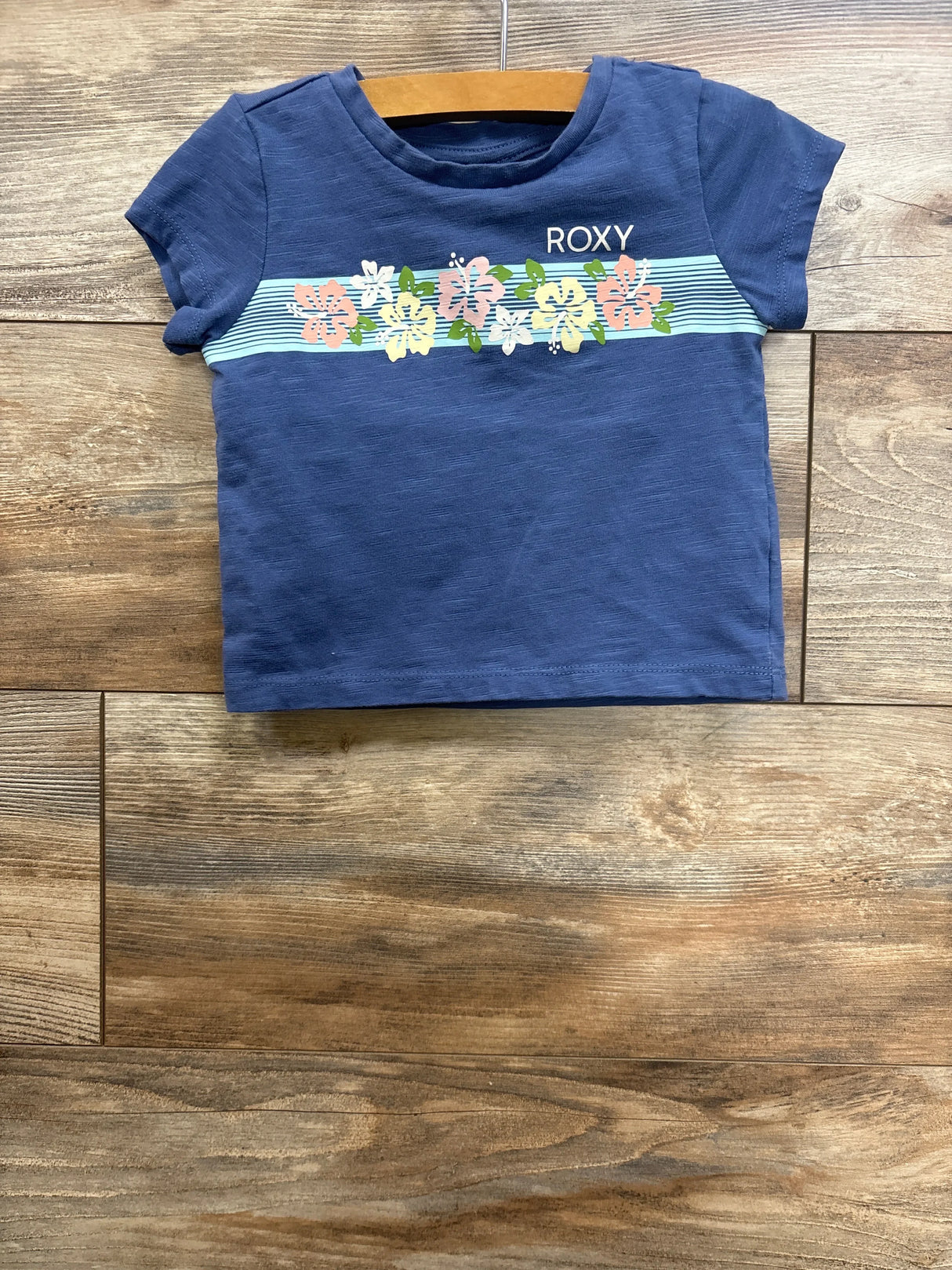 Front flat lay of Roxy toddler short-sleeve shirt size 3T showcasing pastel floral stripe and ROXY logo on wooden floor