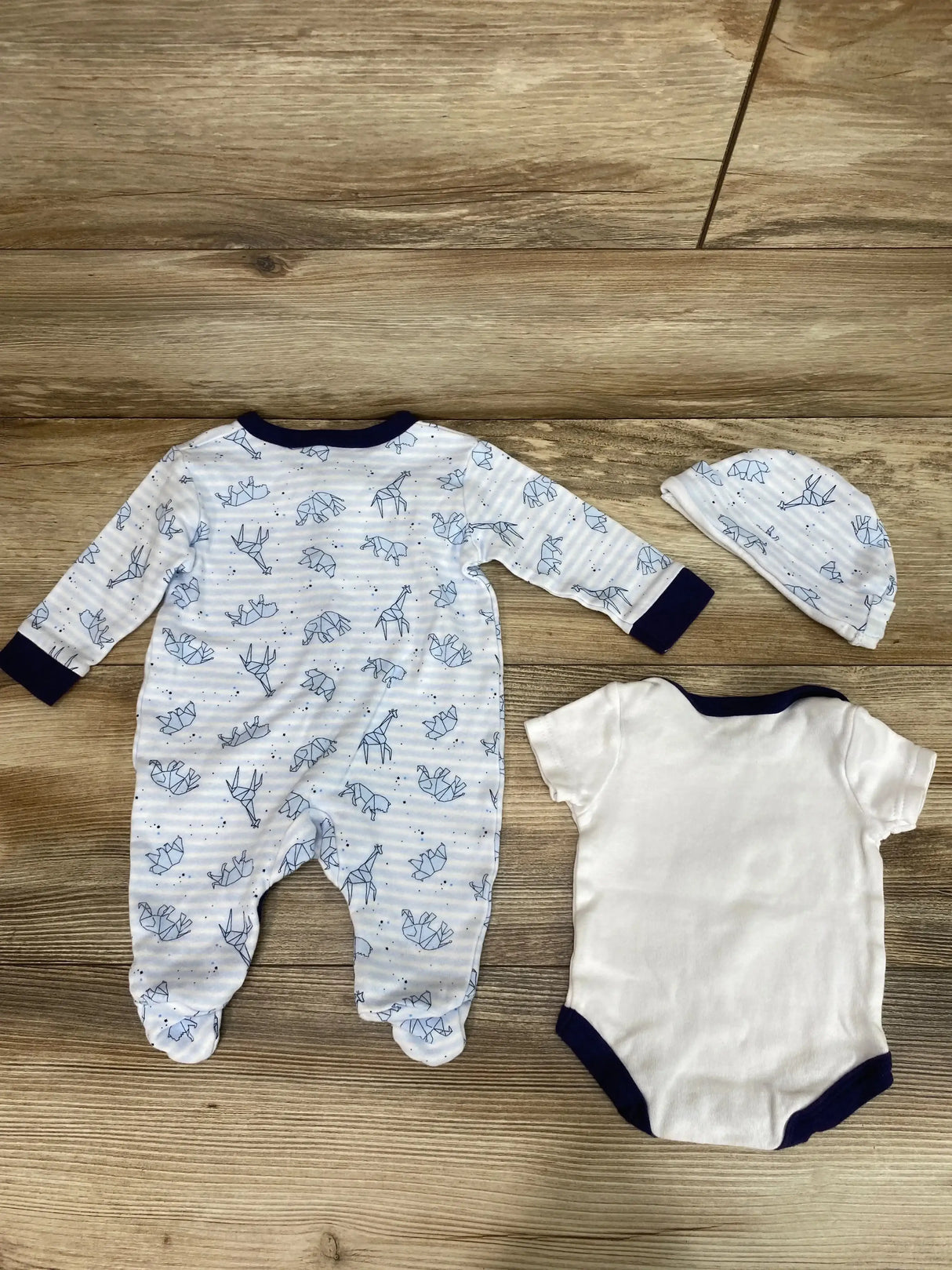 Flat lay of 3-piece baby set: blue animal-print footed sleeper, white bodysuit with navy trim, matching hat on wooden floor