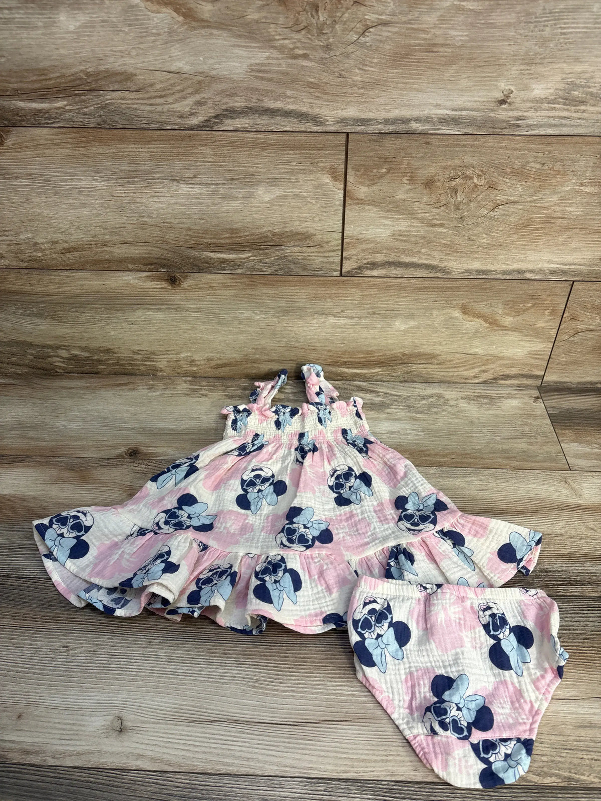 Top view of ivory Disney Baby Minnie Mouse gauze dress and bloomers set with smocked bodice and peplum hem on wood flooring