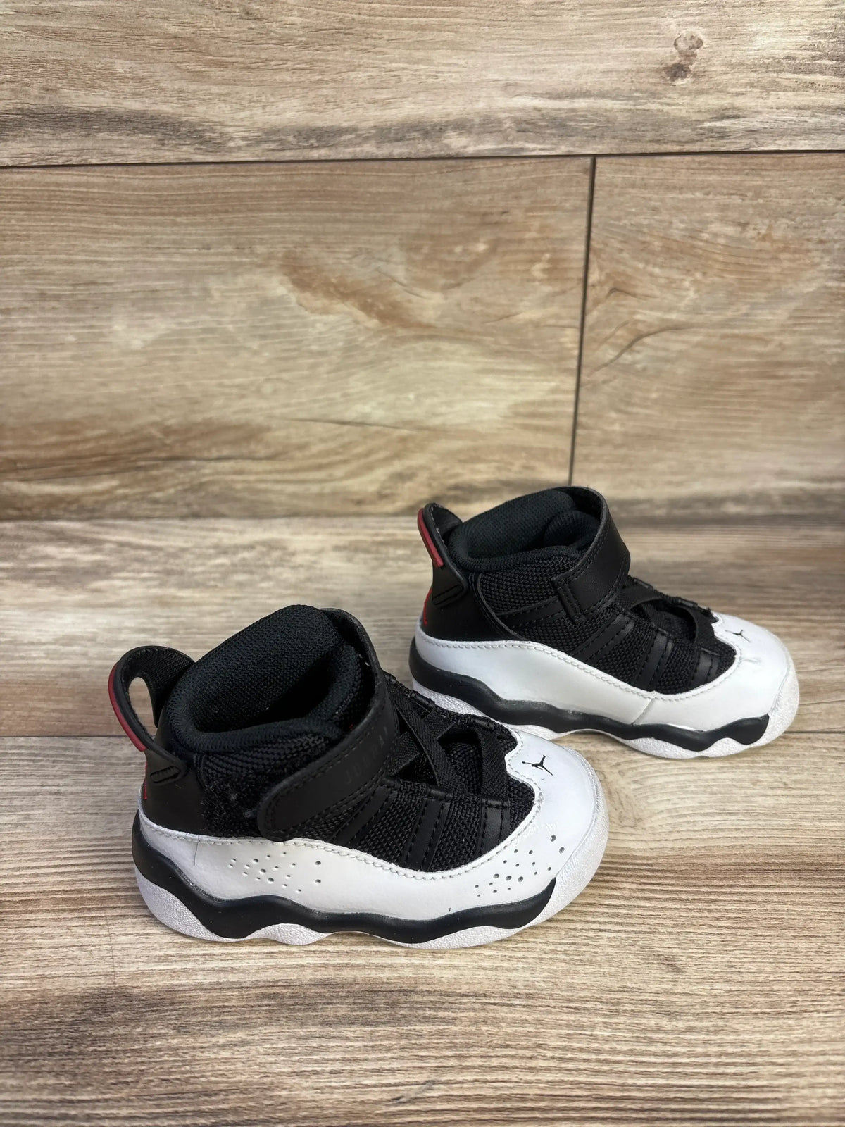 Three-quarter view of Air Jordan 6 Rings toddler sneakers, black and white with Velcro strap and red heel tab on wood floor