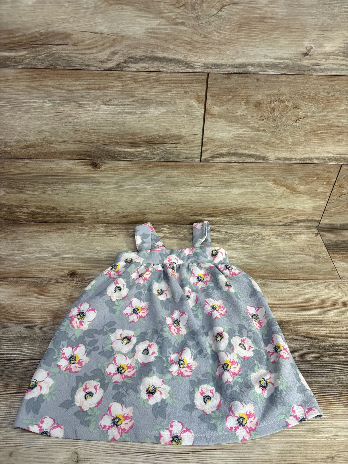 Flat lay of girls' Madison Layne floral dress showcasing grey fabric with white and pink flowers, square straps and gathered waist on wood floor