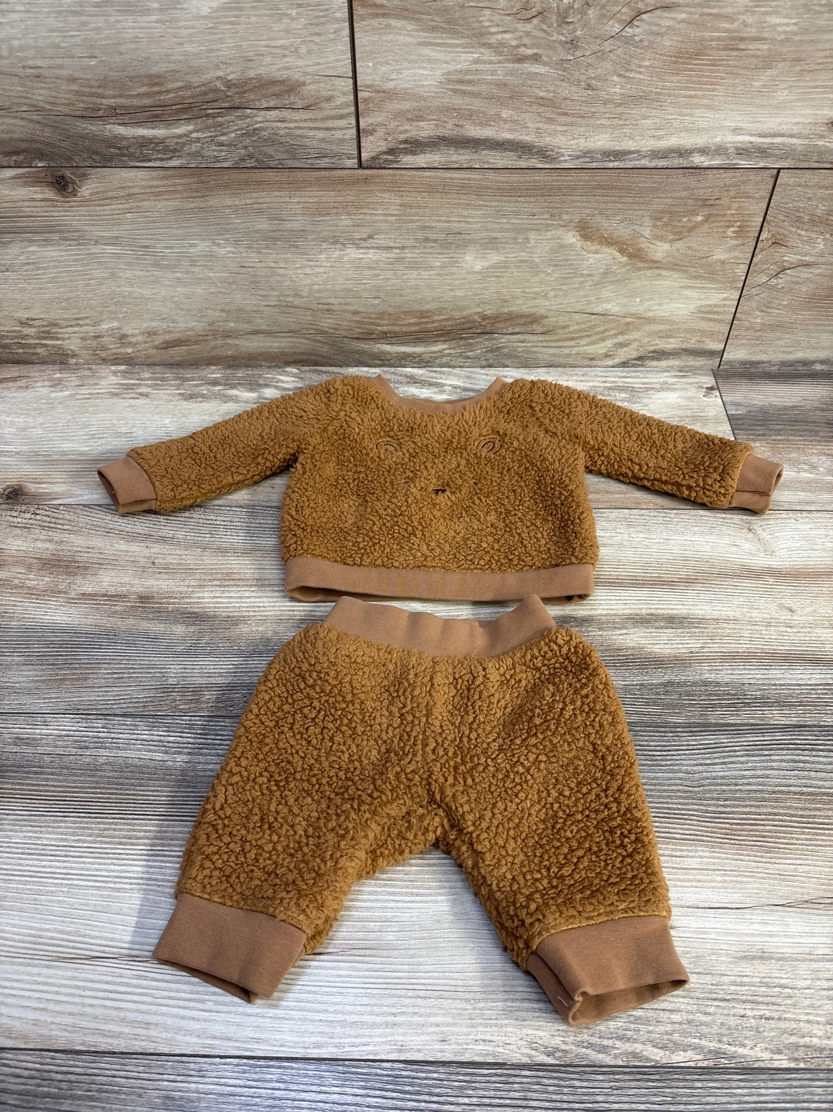 Flat lay of newborn faux-fur sweatshirt and joggers showing brown teddy texture, ribbed cuffs, and bear face on wood background
