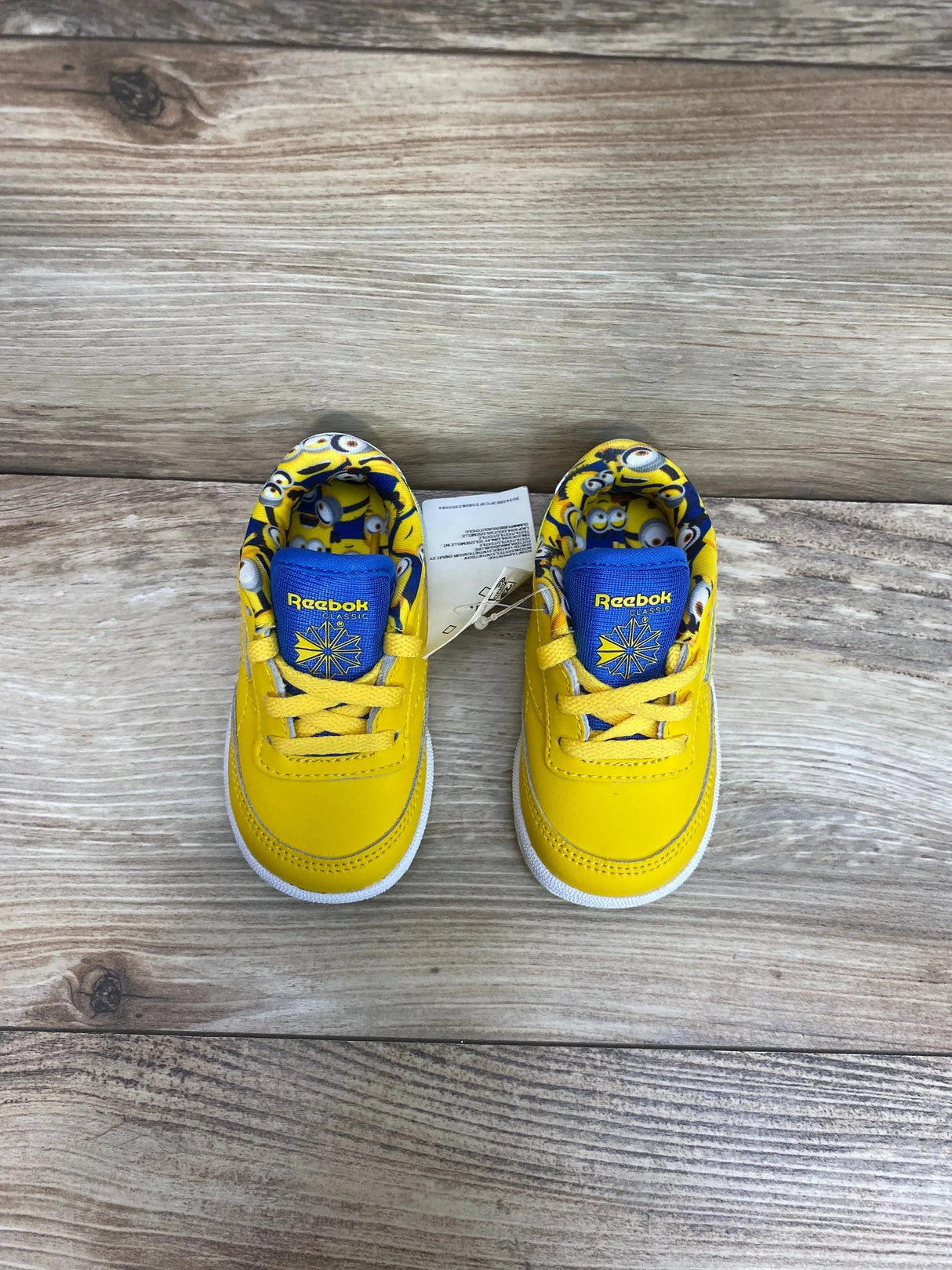 Top view of toddler yellow Reebok Club C 85 sneakers showing blue Reebok tongue and Minions-print lining on wood background
