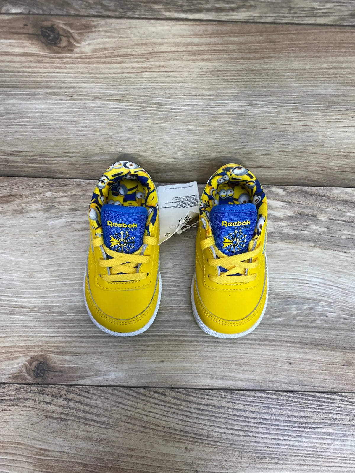 Top view of Reebok Minions Club C 85 toddler sneakers in yellow with blue Minions print tongue and hangtag on wood plank floor