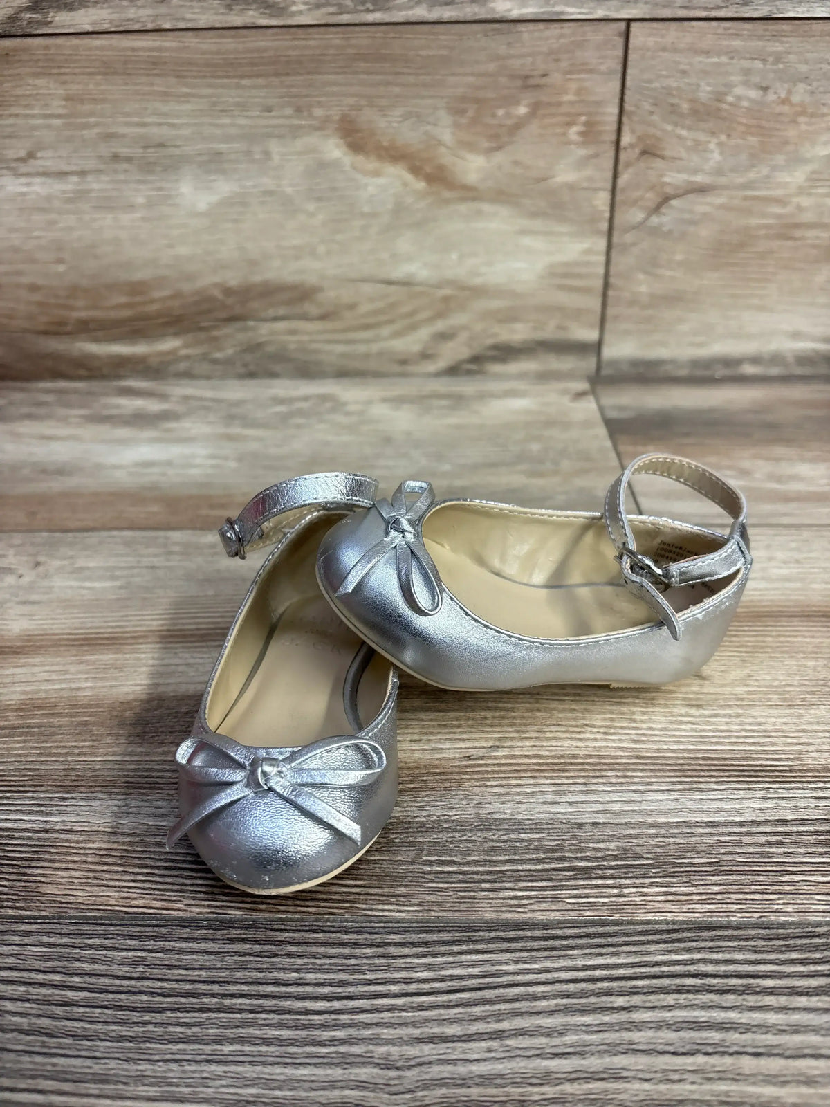 Silver toddler girl shoes with ankle straps and front bows on wooden floor
