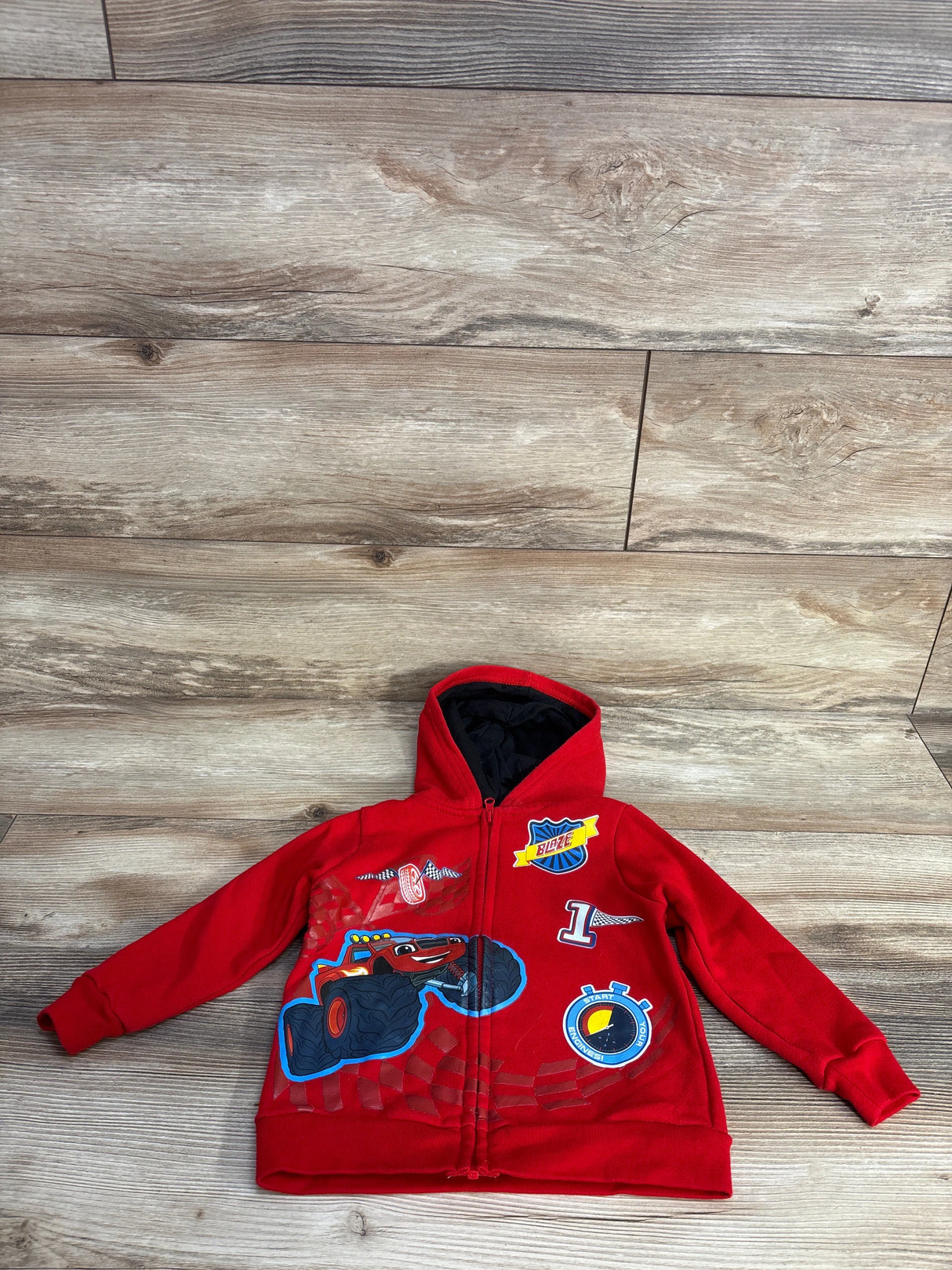 Red kids hoodie with Blaze monster truck design, characters, and racing patches on wooden floor