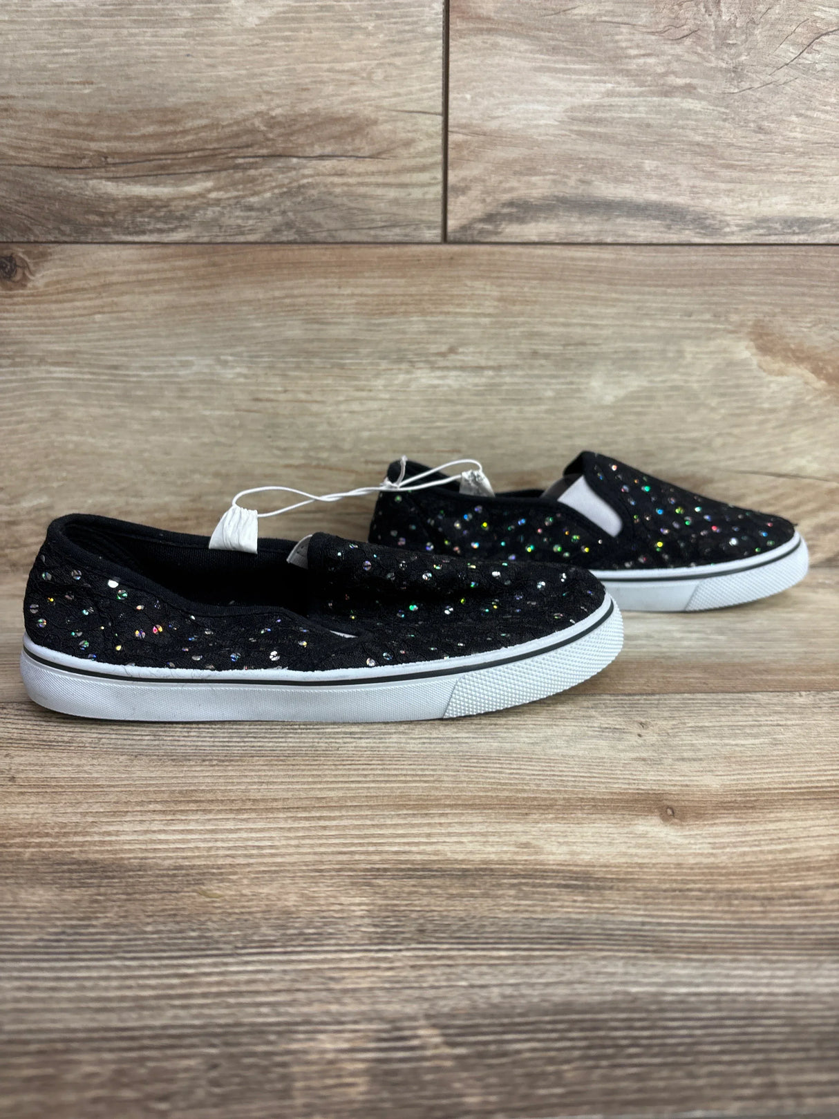Pair of black kids slip-on shoes with colorful glitter dots and white rubber soles on wooden floor