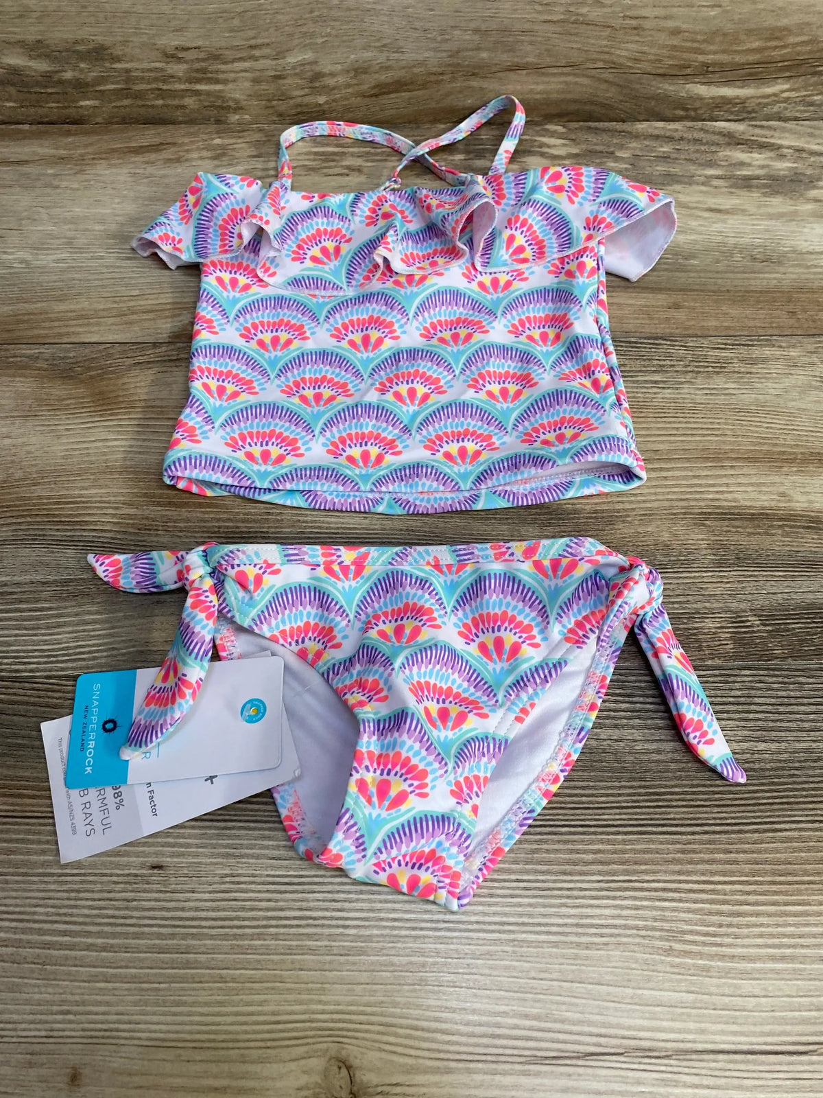 Flat lay of toddler 2T tankini with tags showing multicolor fan-print, ruffle top and side-tie bottoms on wood background