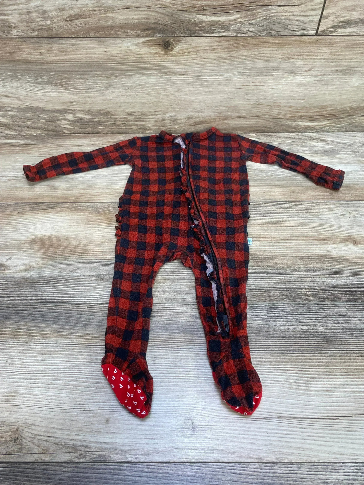 Flat lay of Posh P. buffalo check footie, size 0-3M, showing front zipper with ruffle and red gripper soles on wood