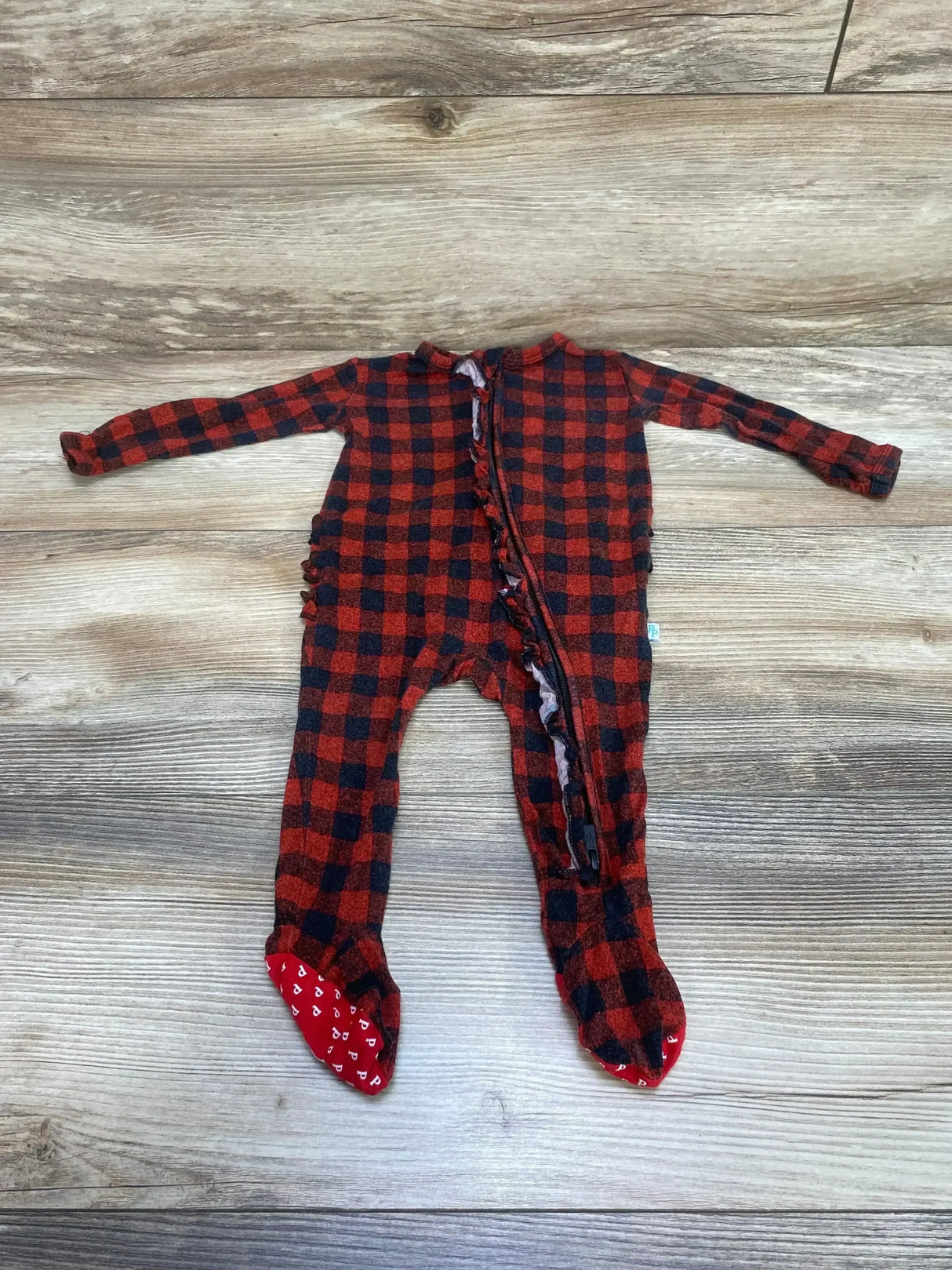 Flat lay of Posh P. buffalo check footie, size 0-3M, showing front zipper with ruffle and red gripper soles on wood
