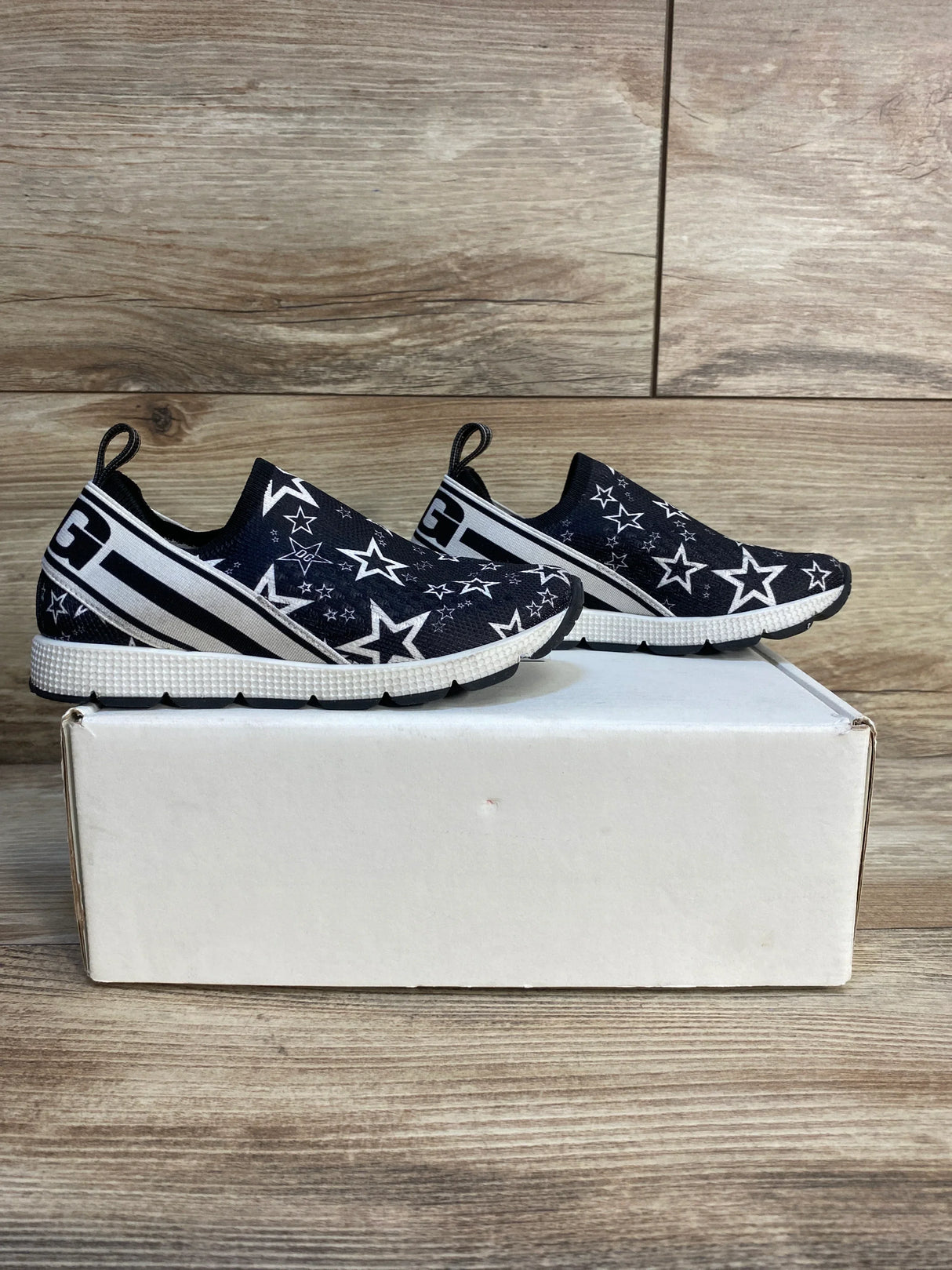 Kids black slip-on sneakers with white star pattern on display box, wooden background