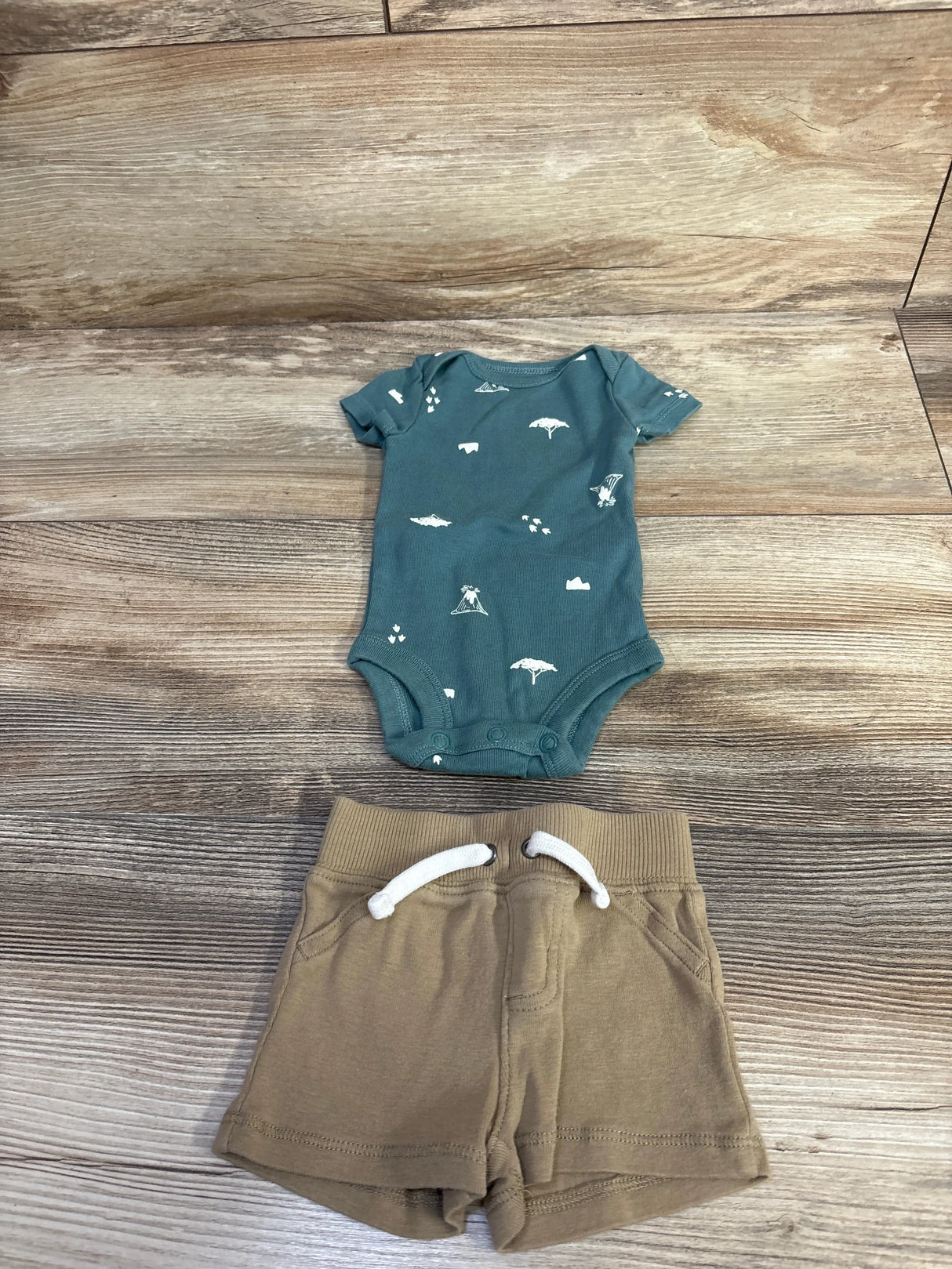 Flat lay of Child of Mine newborn 2-piece set: green bodysuit with white camping icons and tan shorts on wood background
