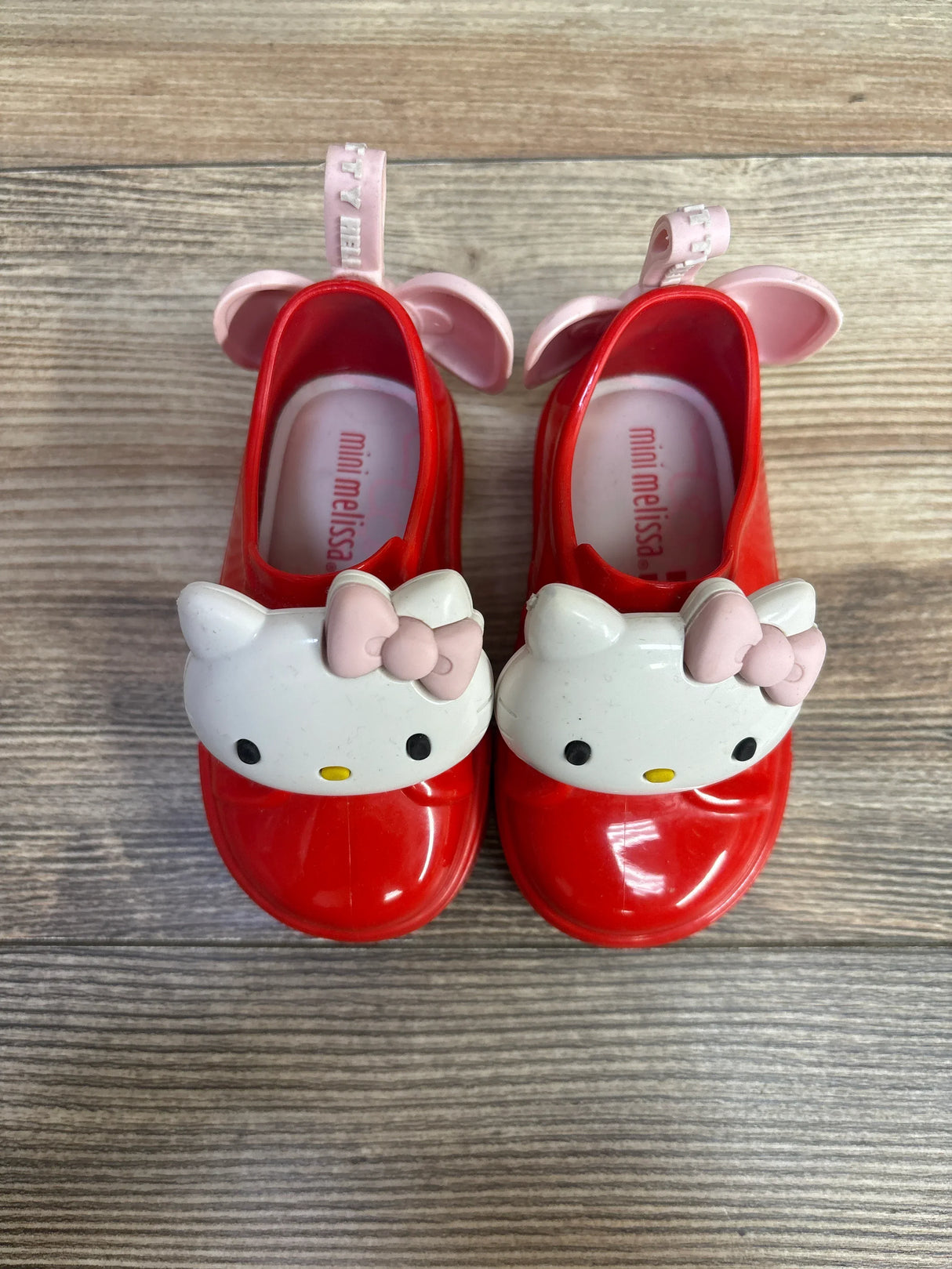 Top view of Mini Melissa Hello Kitty slip-on shoes in red showcasing kitty face appliqué with pink bow on wood plank floor