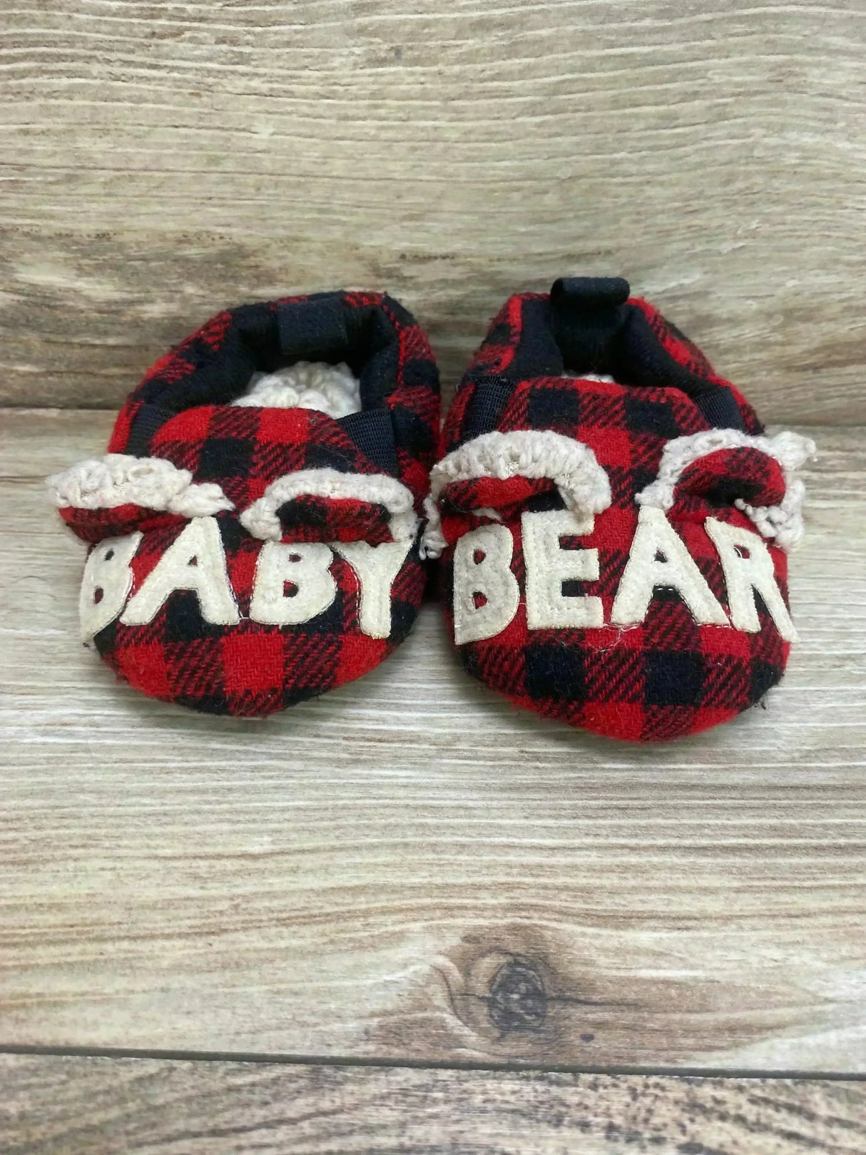 Front view of red and black buffalo plaid baby slippers with white ‘BABY BEAR’ letters and faux-shearling ears on light wood background