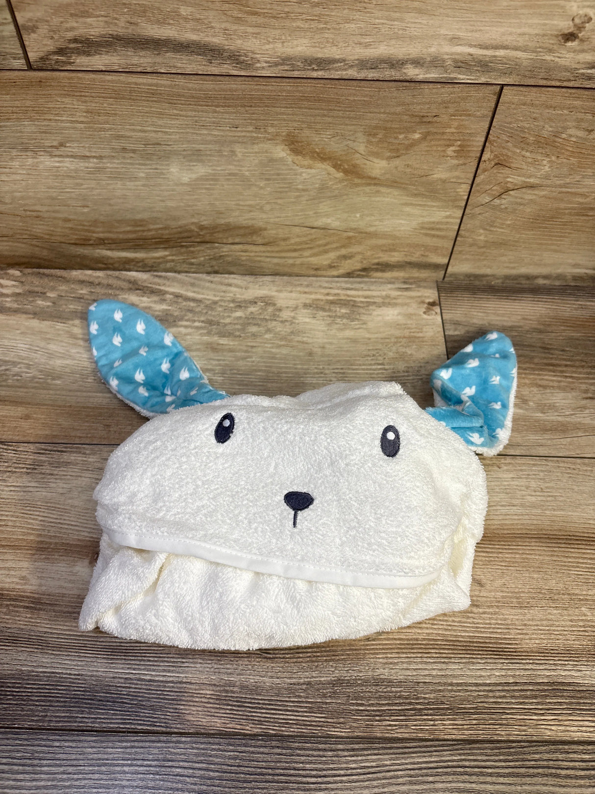 Top view of white hooded bunny baby towel showing embroidered face and blue dove patterned ear lining on wooden floor