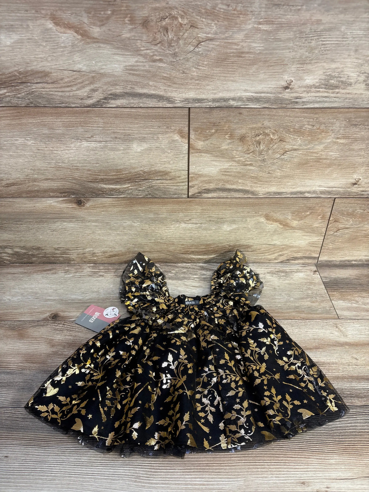 Front view of Modern Moments x Wicked toddler dress with black and gold floral pattern and ruffle sleeves on wood floor