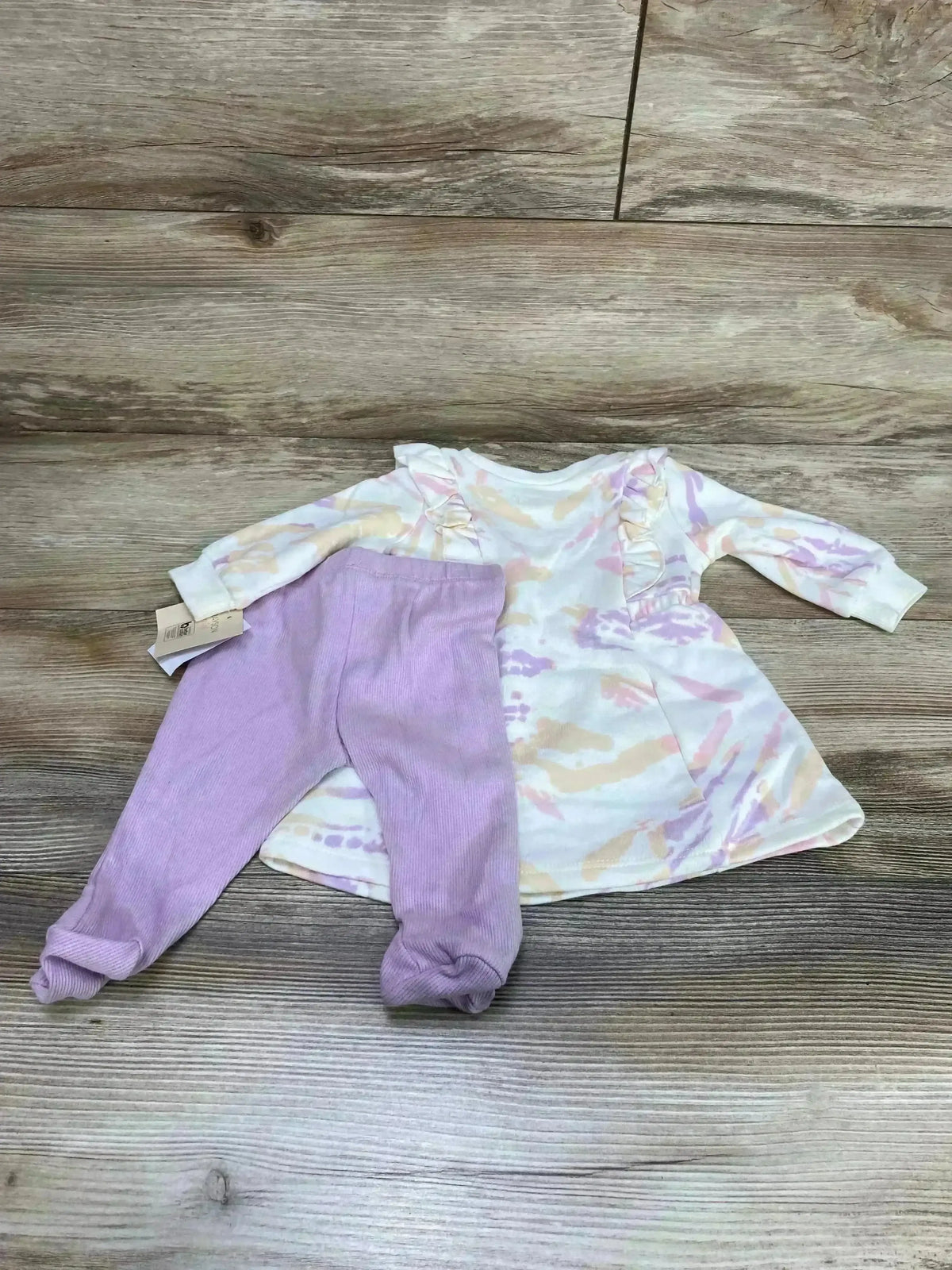 Top-down view of Grayson Mini 2-piece tie-dye dress with ruffled shoulders and lavender footed pants, size 0-3M on wood floor