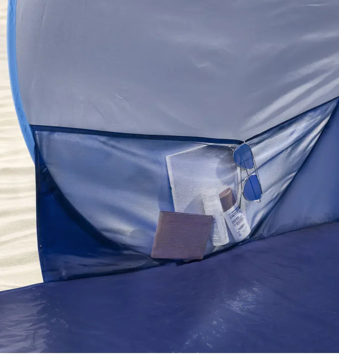 Blue beach tent interior with mesh pocket holding sunglasses, sunscreen, and a book on sand