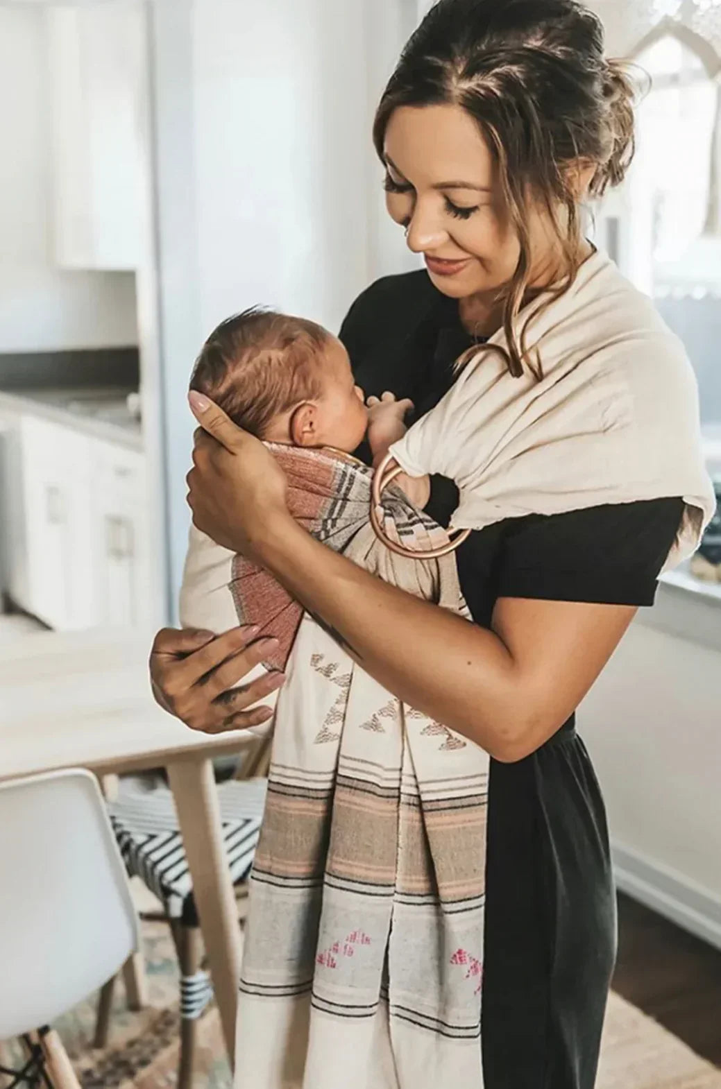 Three-quarter view of Beachwood Aztec handwoven baby ring sling (8-35 lb) showing patterned fabric and wooden rings, worn in modern kitchen